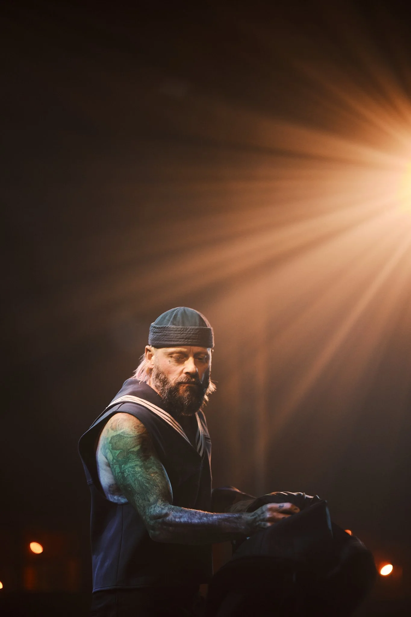 A man with a beard and tattoos on his arms, wearing a beanie and a sleeveless shirt, looks down while holding a black bag. Bright light from the right illuminates the scene, creating a warm glow.