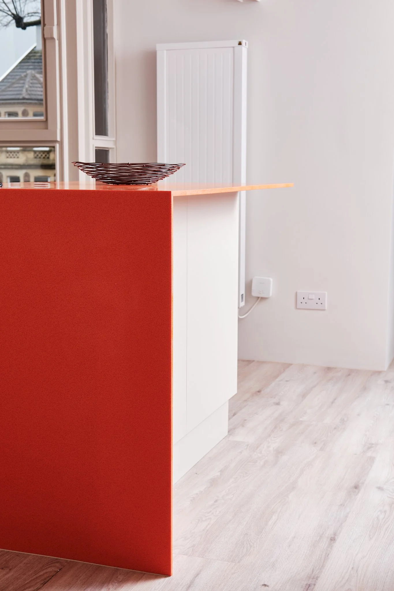 Part of a modern kitchen with a white cabinet, a black wire fruit bowl on an orange-colored breakfast bar, a window with a view of neighboring buildings, and a white radiator on the wall.