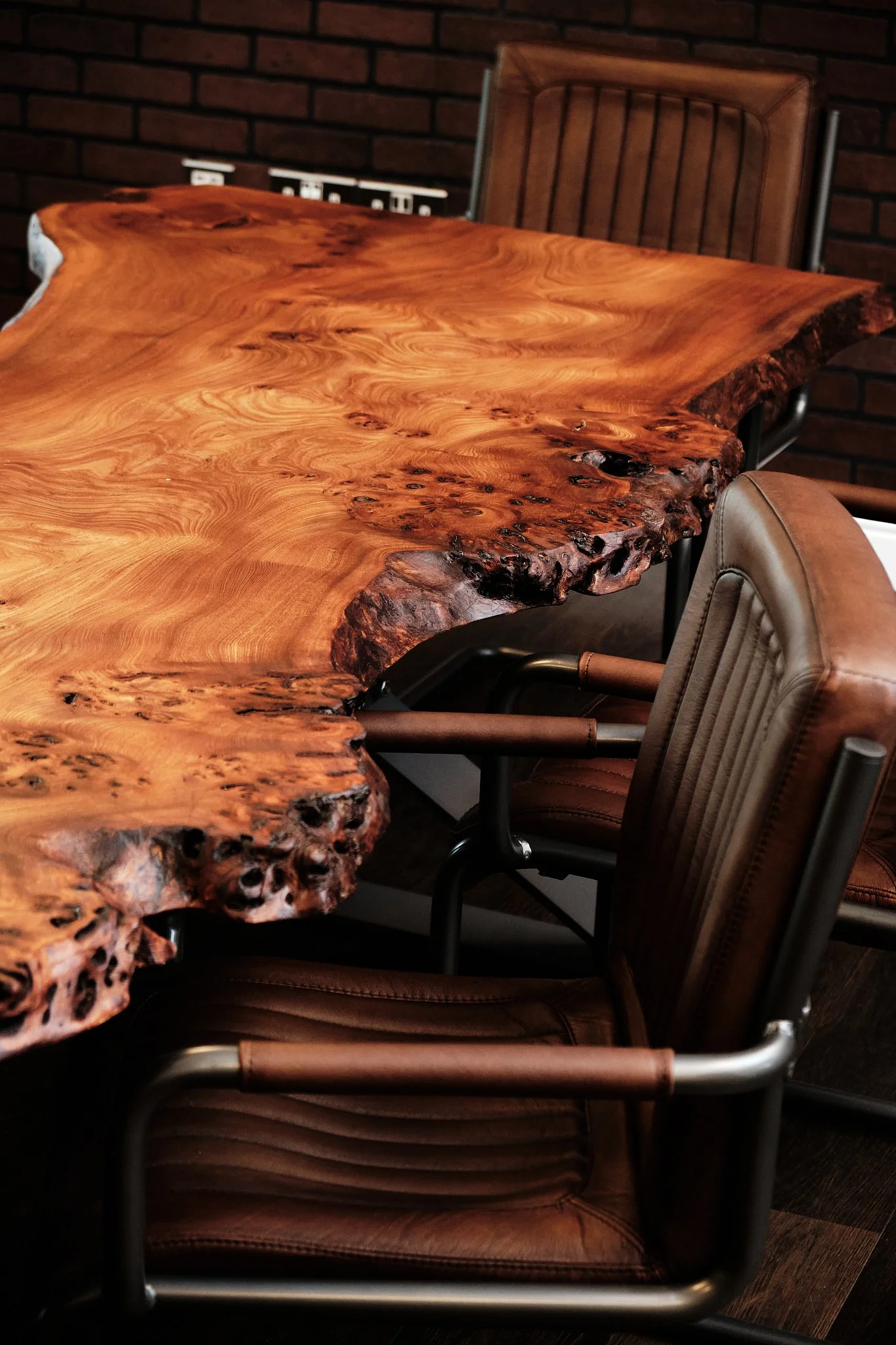 A wooden conference table with a natural, uneven edge in a room with brick walls and leather chairs around it.