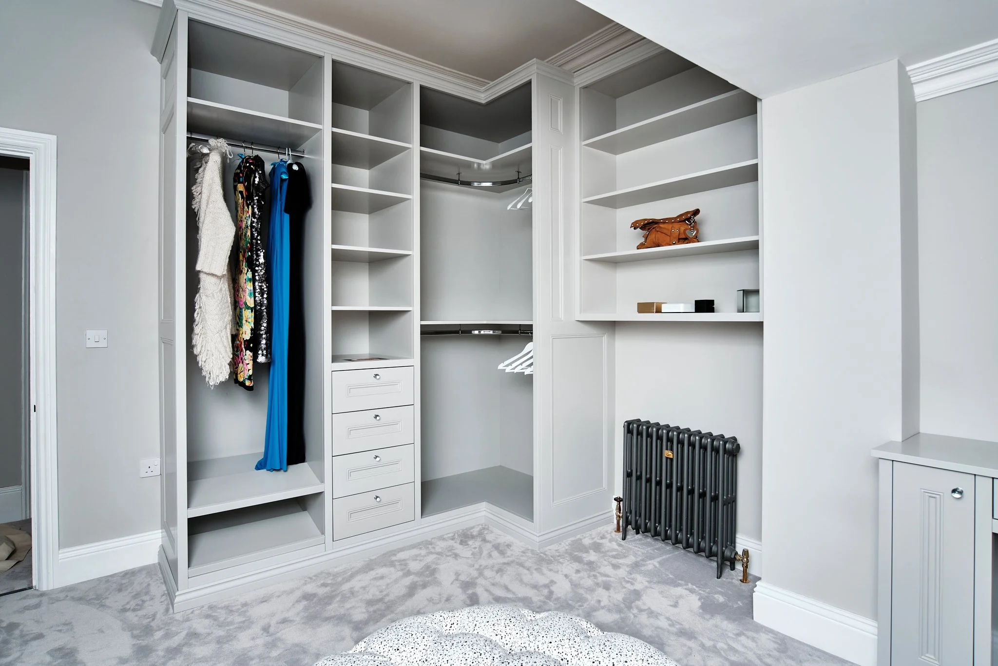 Empty built-in closet with clothes on a rail, shelves, drawers, and a small cabinet, in a modern bedroom with light gray walls and carpet