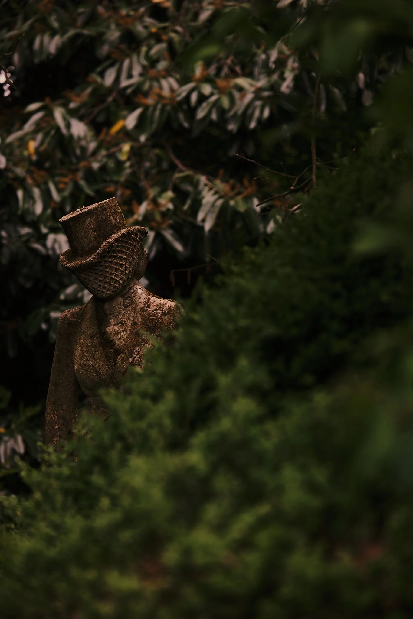 Rusty garden statue of a woman wearing a top hat and a textured scarf, partially hidden among green bushes and dark leafy plants. Hotel interior photography at THE PIG-in the Cotswolds
