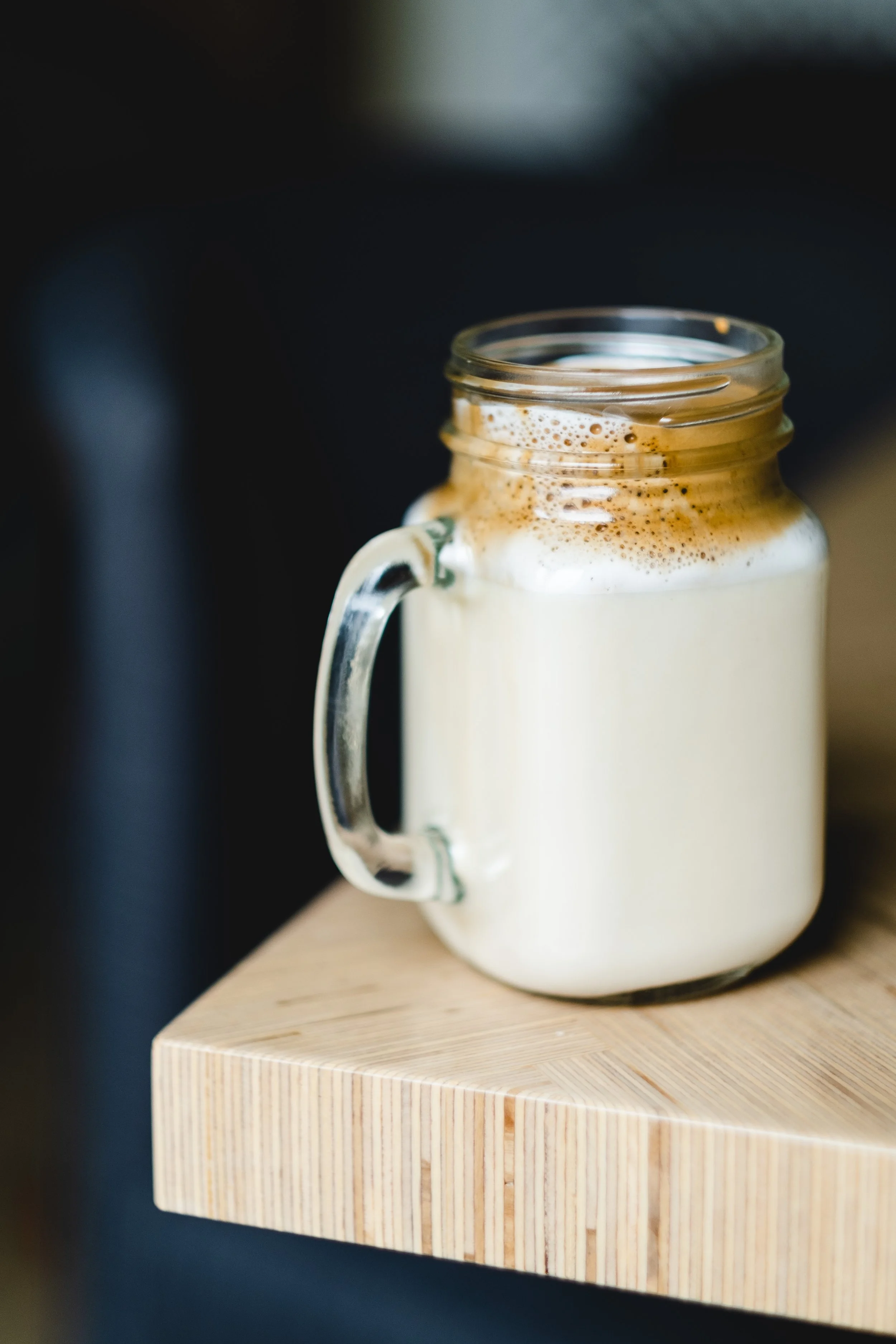 A glass mason jar mug filled with a frothy, layered coffee drink, placed on a wooden surface.