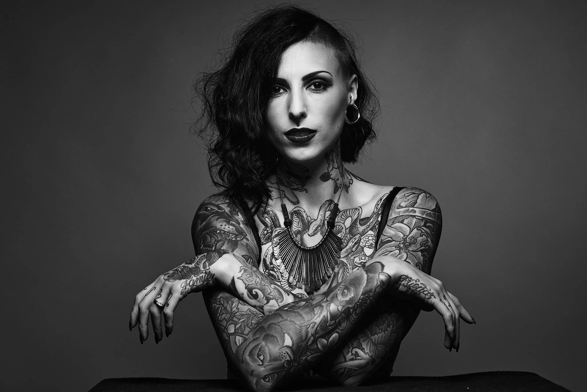 Black and white photo of a woman with tattoos on her arms and chest, wearing earrings and a bold necklace, with short wavy hair, looking directly at the camera.