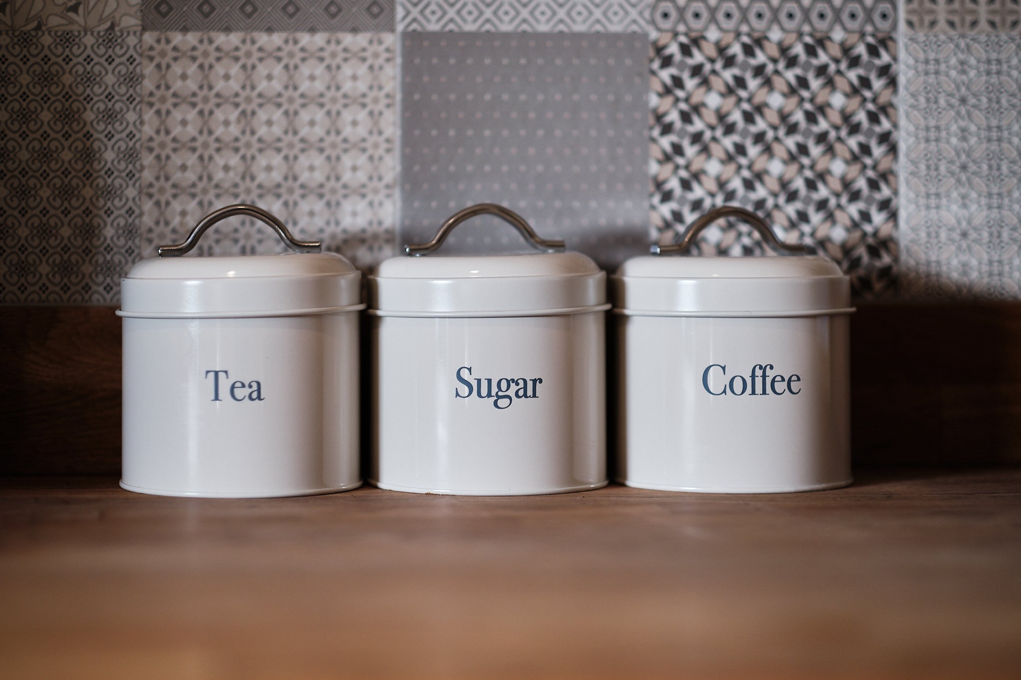 Three white metal canisters labeled Tea, Sugar, and Coffee on a wooden surface with patterned tiles in the background.