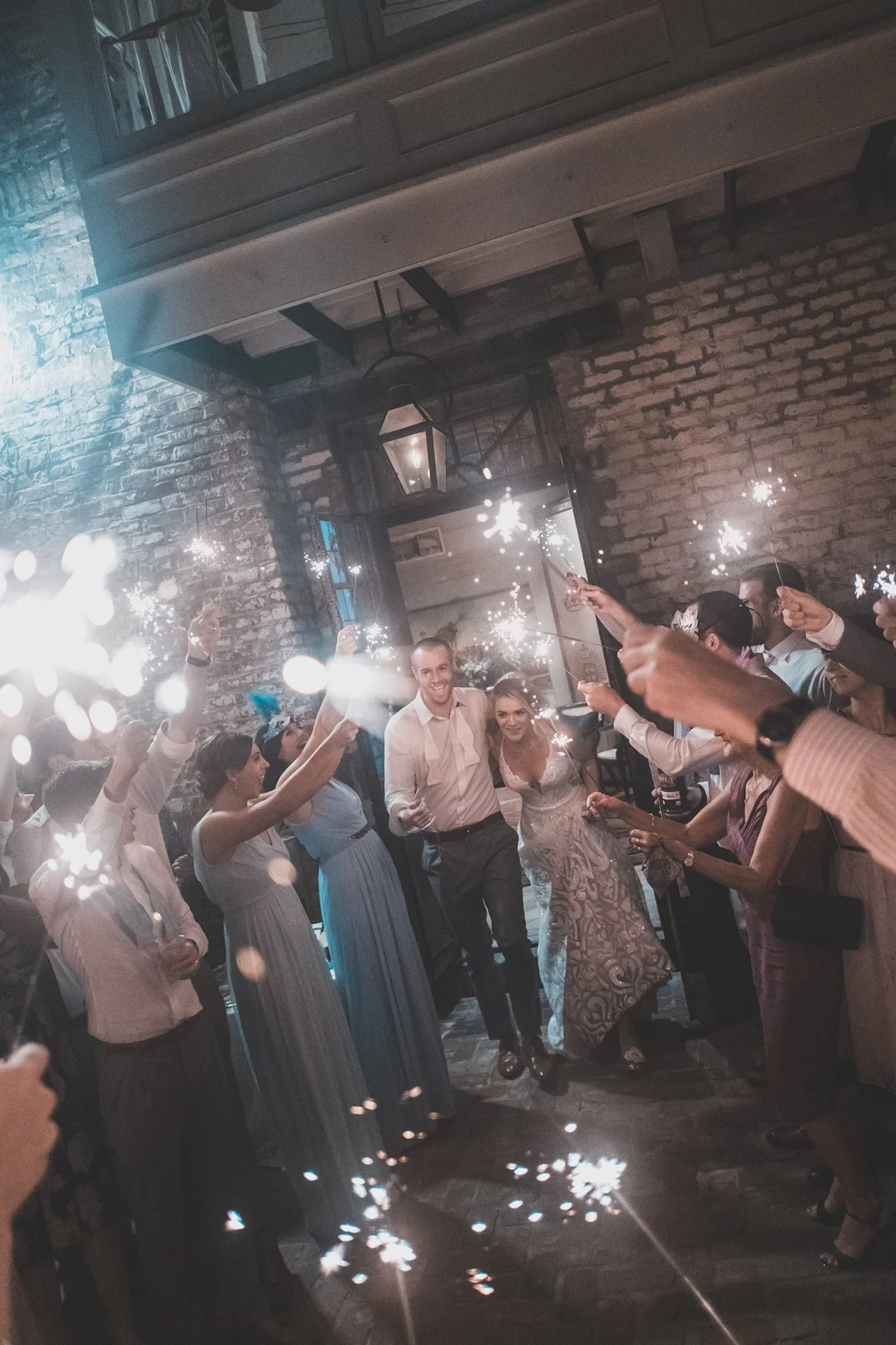 People celebrating with sparklers indoors, smiling and raising their hands, with a rustic brick wall and hanging lanterns in the background.