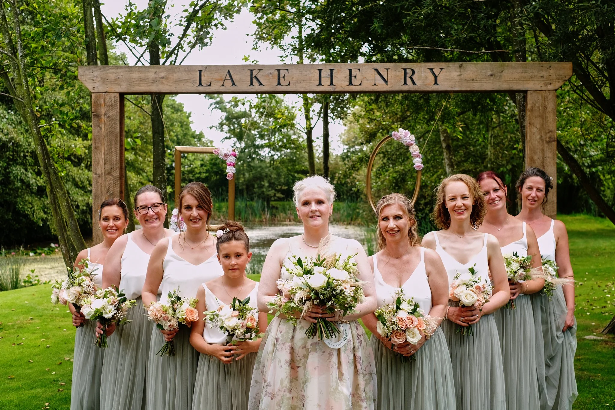 A group of women and girls dressed in white tops and long gray skirts, holding bouquets of flowers, standing in front of a wooden sign that reads 'Lake Henry' during a wedding celebration.