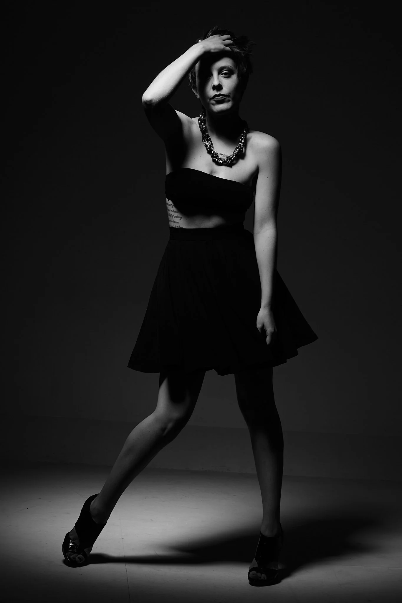 A woman in a black dress and high heels standing against a dark background, posed with one hand on her head and the other hanging down, wearing a necklace.