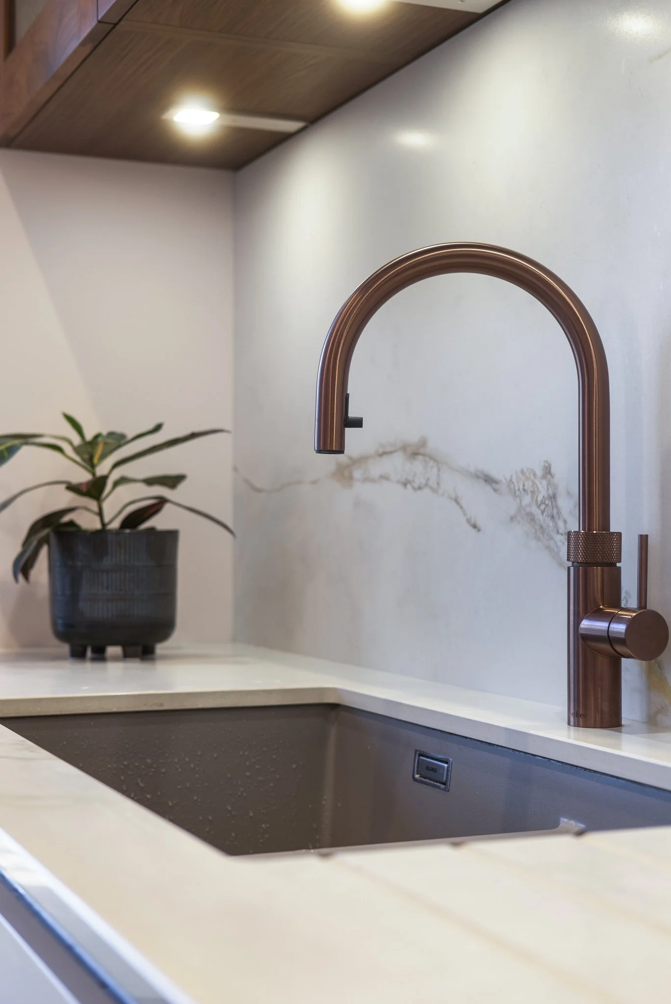 Modern kitchen sink with a copper faucet, white marble countertop, and a potted plant, with wooden cabinetry above and recessed lighting.