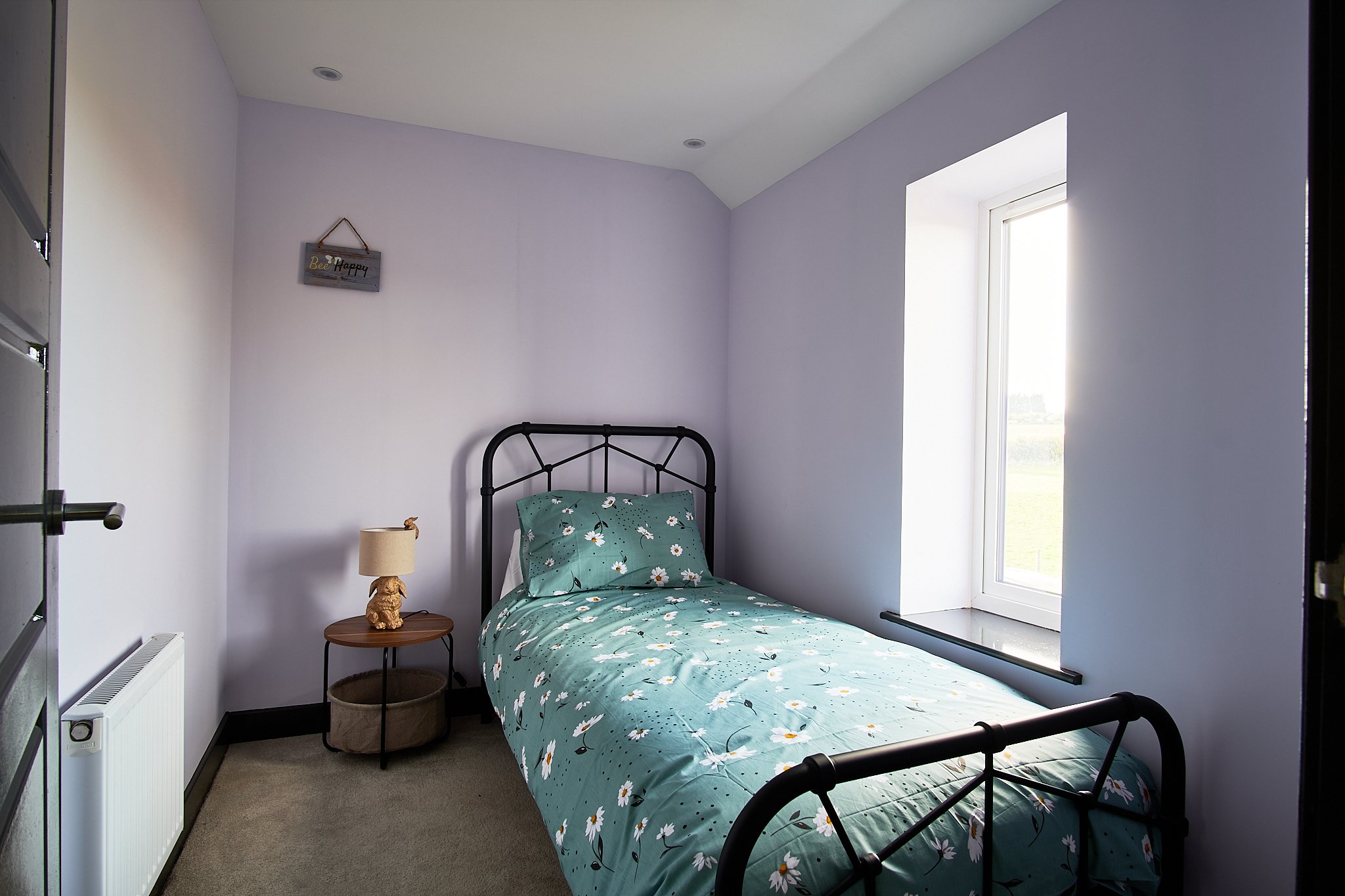 A small bedroom with a black metal bed frame, floral bedding, a side table with a lamp, and a window with a view of the outdoors.
