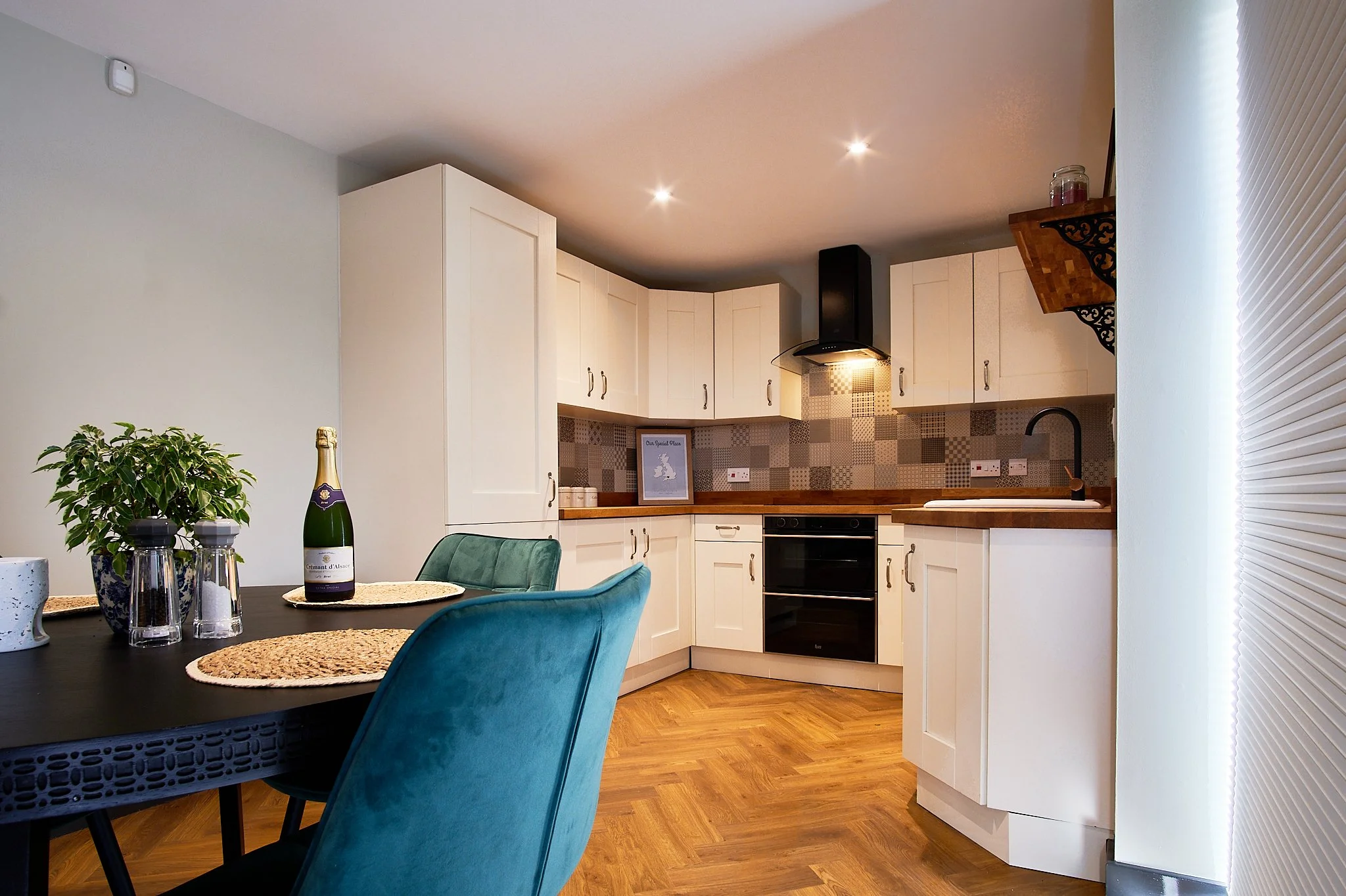 Modern kitchen with white cabinets, wooden countertops, black oven, and patterned tile backsplash. Dining table with teal and green chairs, potted plant, and a bottle of sparkling wine.