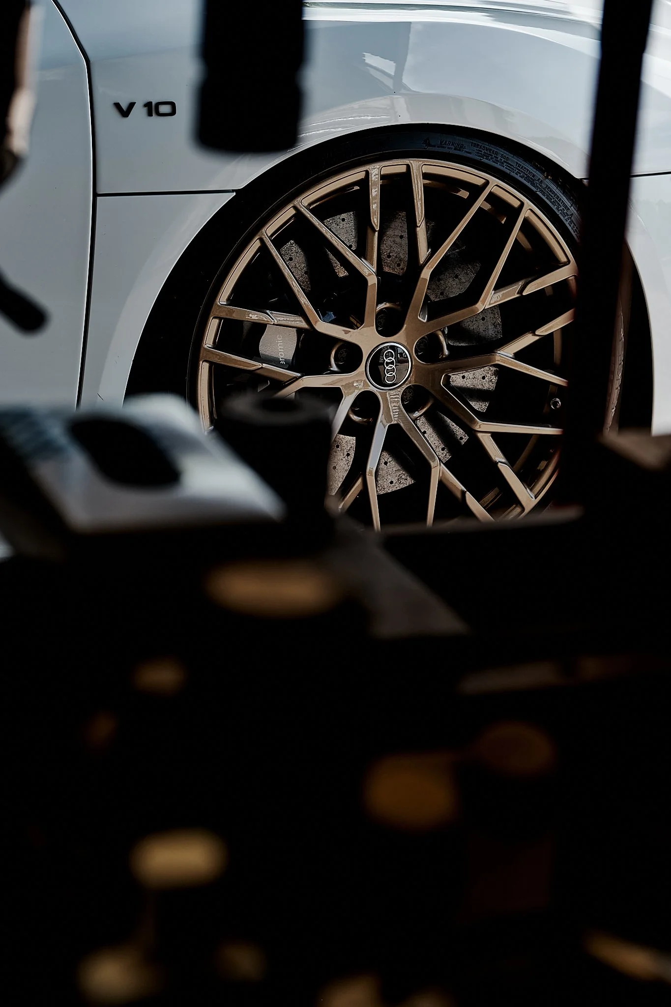 Close-up of a white Audi car with a gold-colored alloy wheel and black brake caliper, seen through a dark frame.