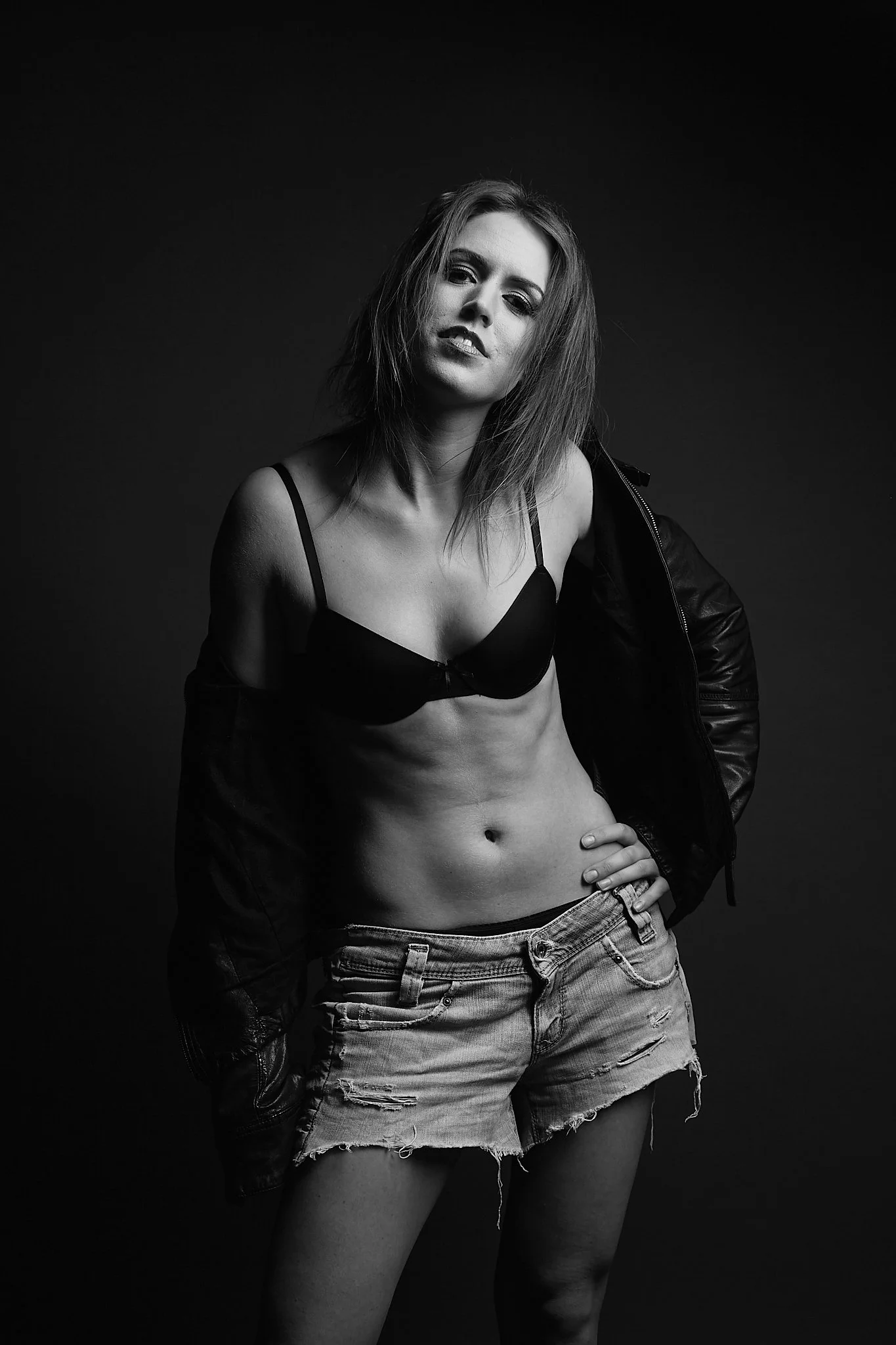 A black-and-white photo of a young woman with shoulder-length hair, wearing a black bra, ripped denim shorts, and a black jacket draped over her shoulders. She is standing against a dark background, with one hand on her hip and a confident expression