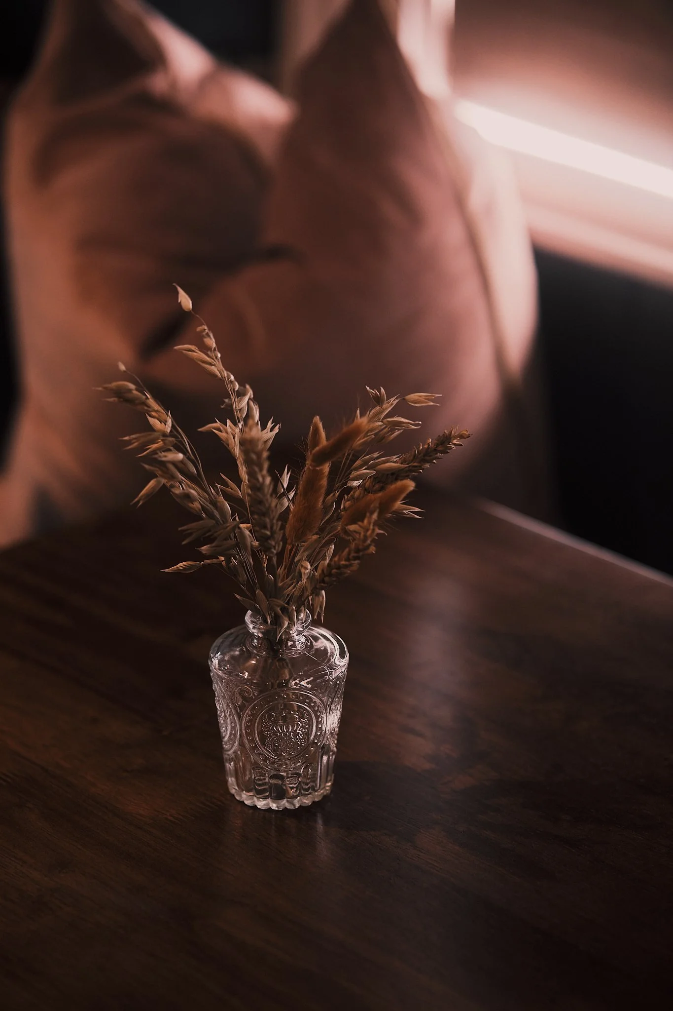 A glass vase with dried wheat and grass stems on a wooden table in dim, warm lighting with a blurred background of a pinkish-brown cushion and bedding.