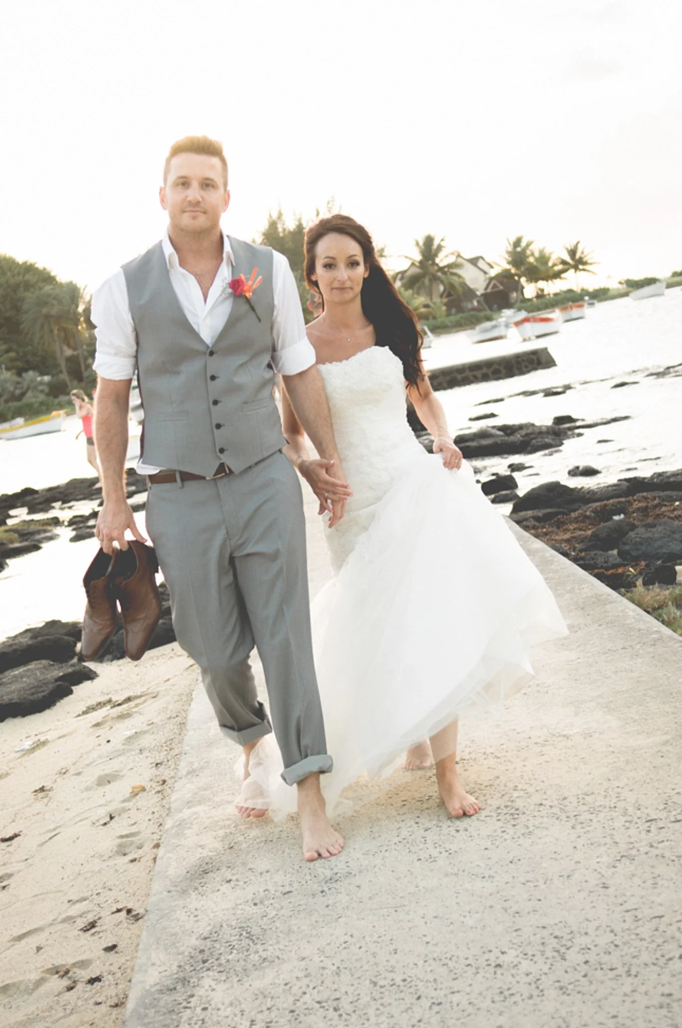 A bride and groom walking barefoot on a sandy path near the ocean, holding hands, with boats and palm trees in the background, during sunset.