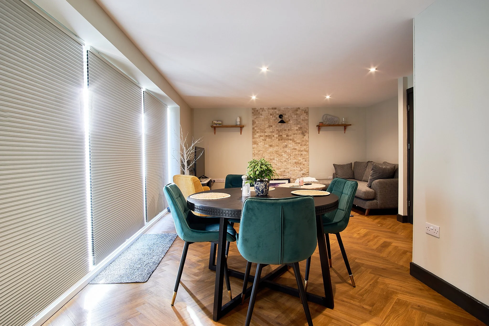 Living and dining area with a round table surrounded by teal and yellow chairs, a stone feature wall, a gray sofa, and large windows with closed blinds.
