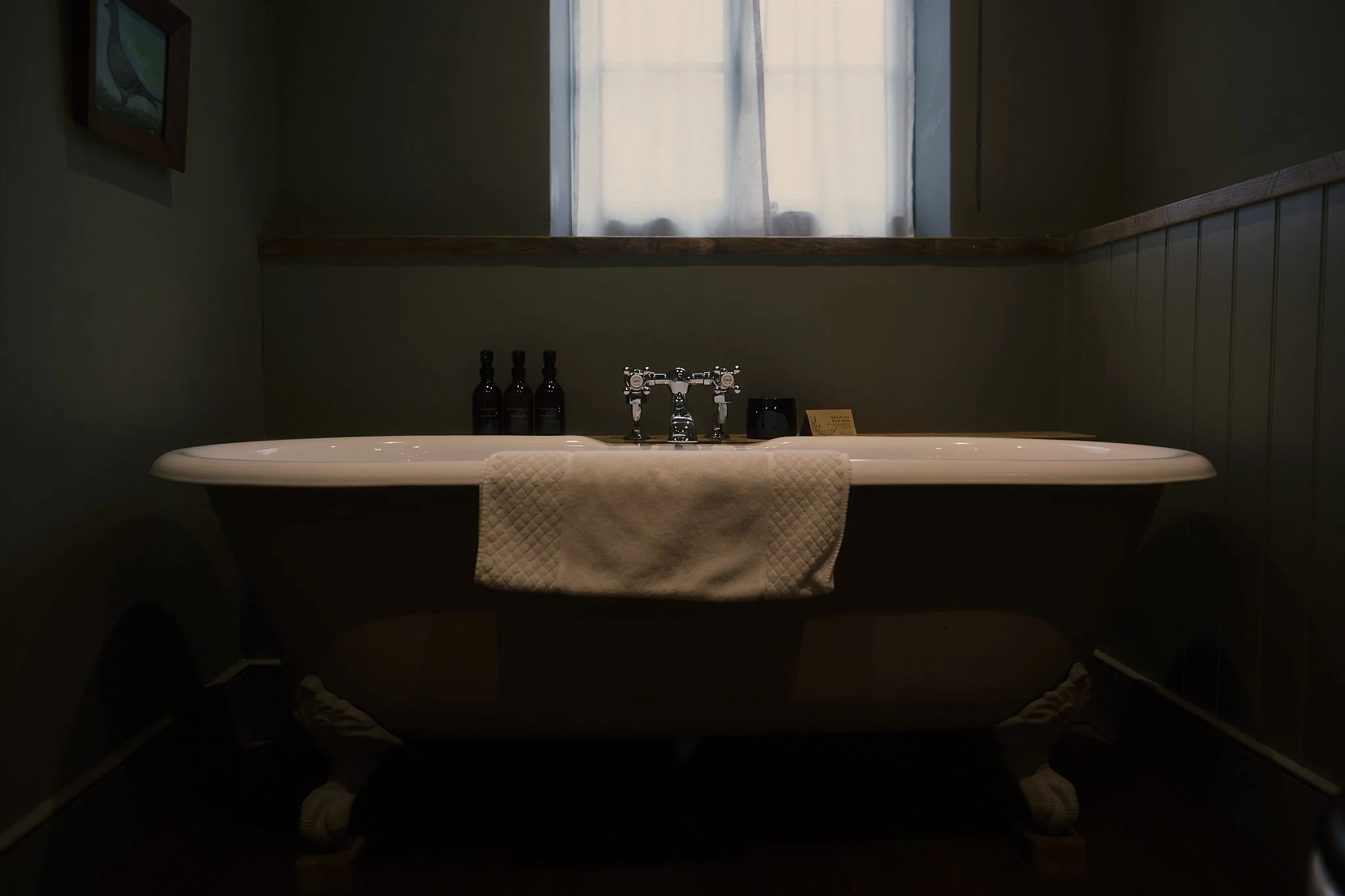 Vintage clawfoot bathtub under a window with sheer curtains, with three dark bottles on the ledge. Hotel interior photography at THE PIG-in the Cotswolds