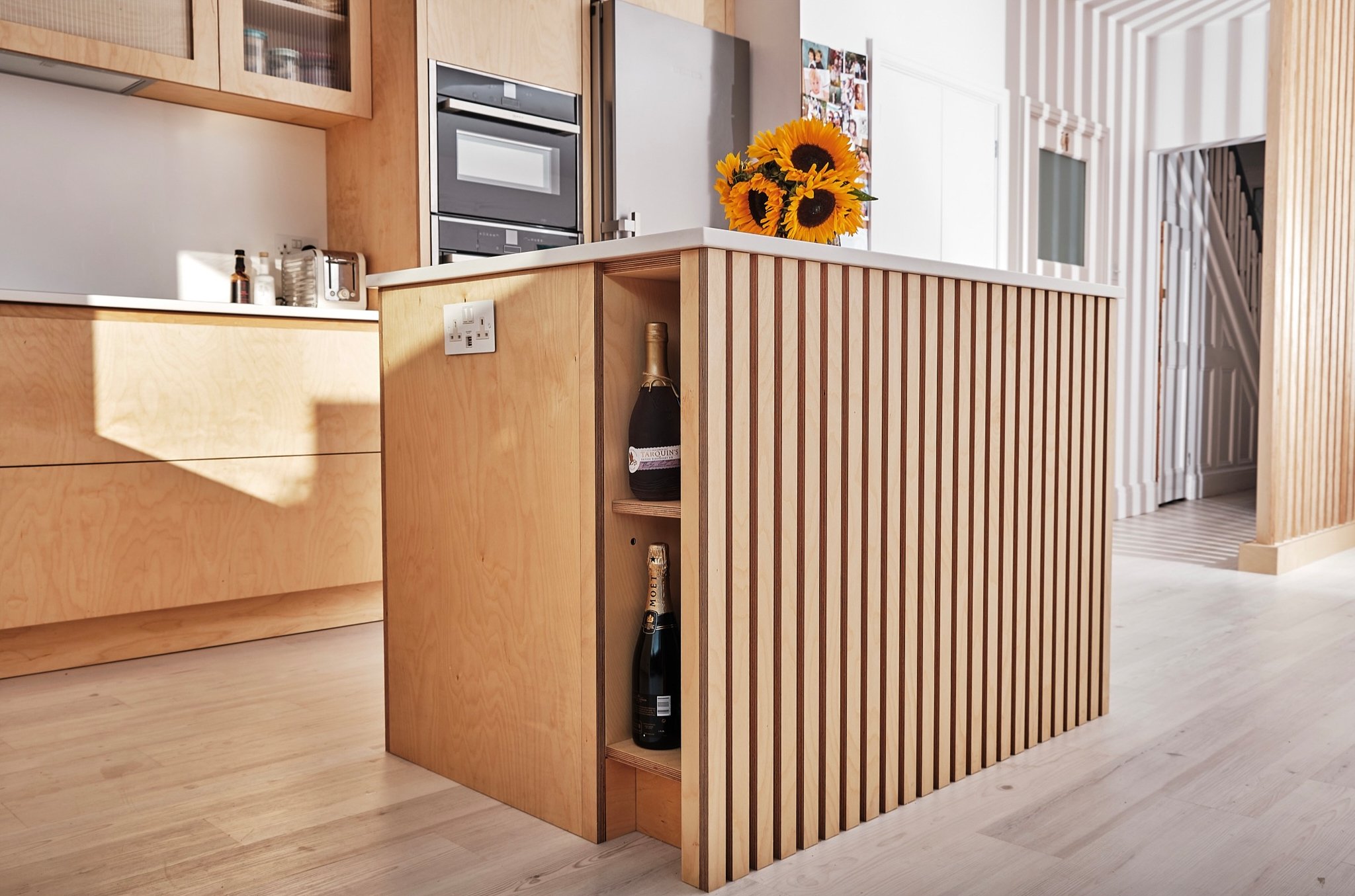 Modern kitchen with light wood cabinetry and a kitchen island holding a bouquet of sunflowers, with wine bottles stored in a side shelf.