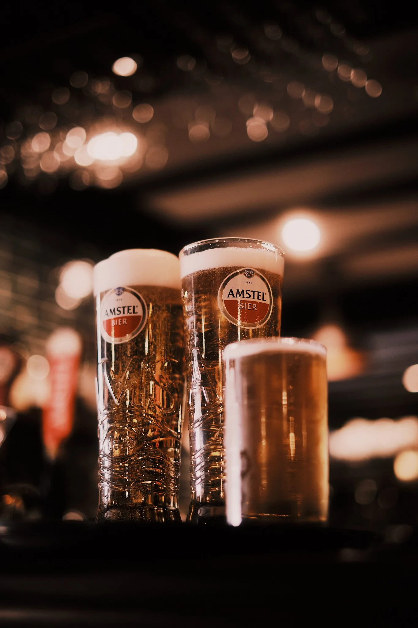 Three glasses of beer with foam on top, placed on a bar or table, with warm lighting and a blurred background.