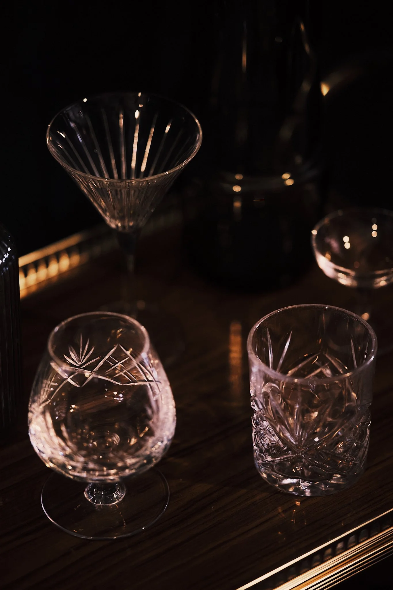 Several clear glassware pieces with intricate cut patterns, including a martini glass, a whiskey glass, and a short tumbler, arranged on a dark wooden surface with a dimly lit background.