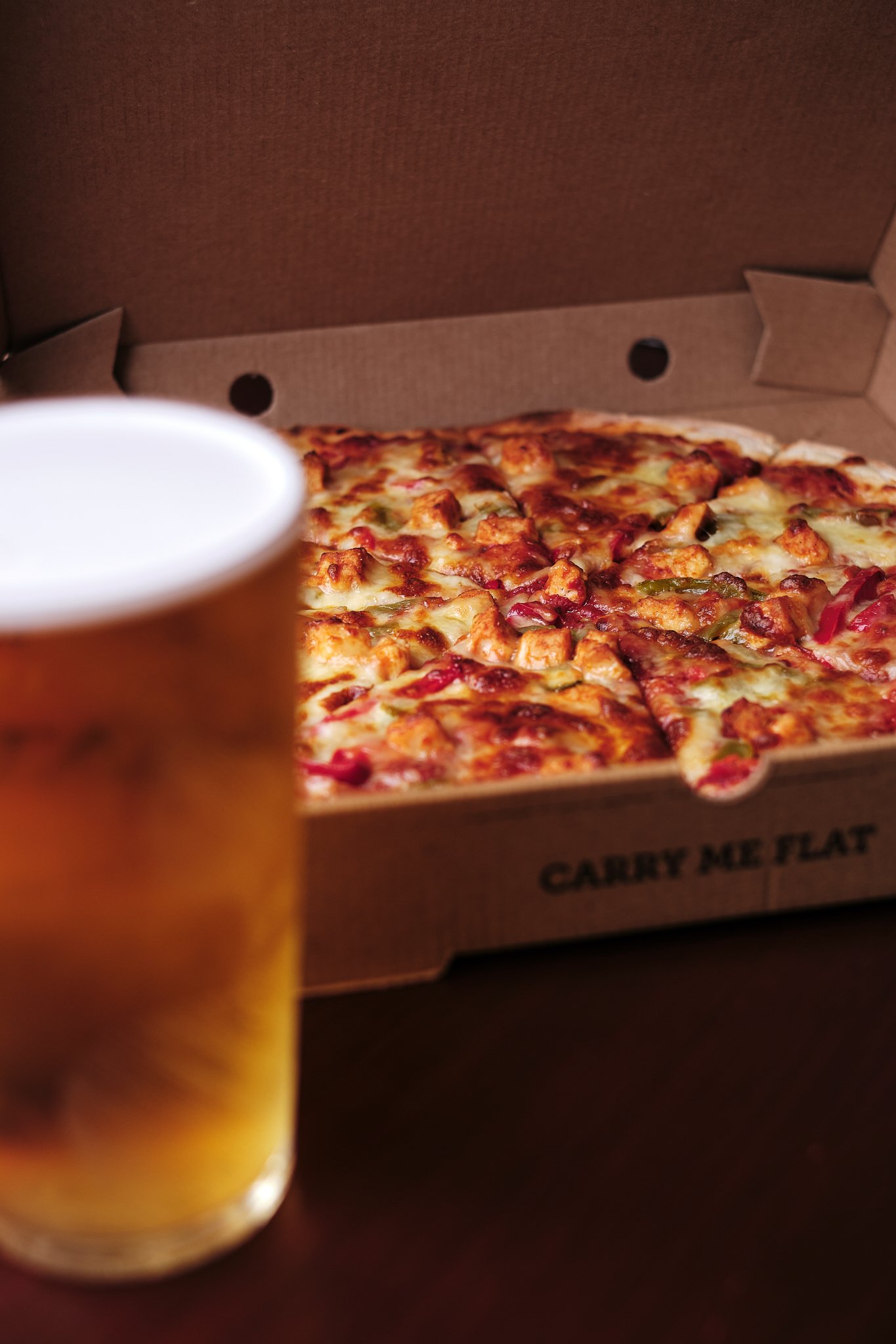 A partially visible glass of beer in front of a box of pizza with cheese, chicken, and vegetables.