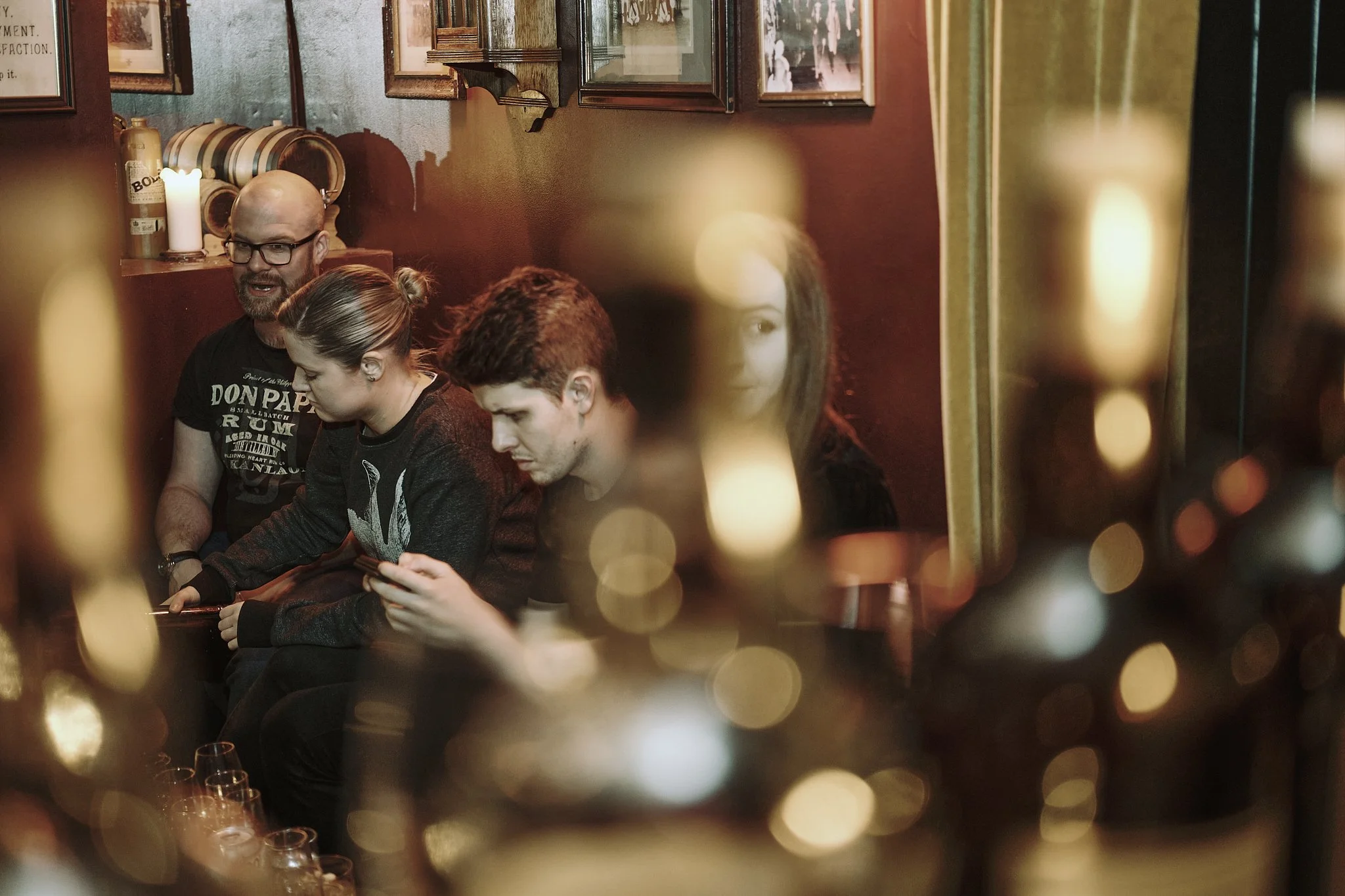 Four people sitting in a dimly lit bar, two men and two women, with some overlooking wine glasses in the foreground, framed by the blurry shape of bottles and glasses.