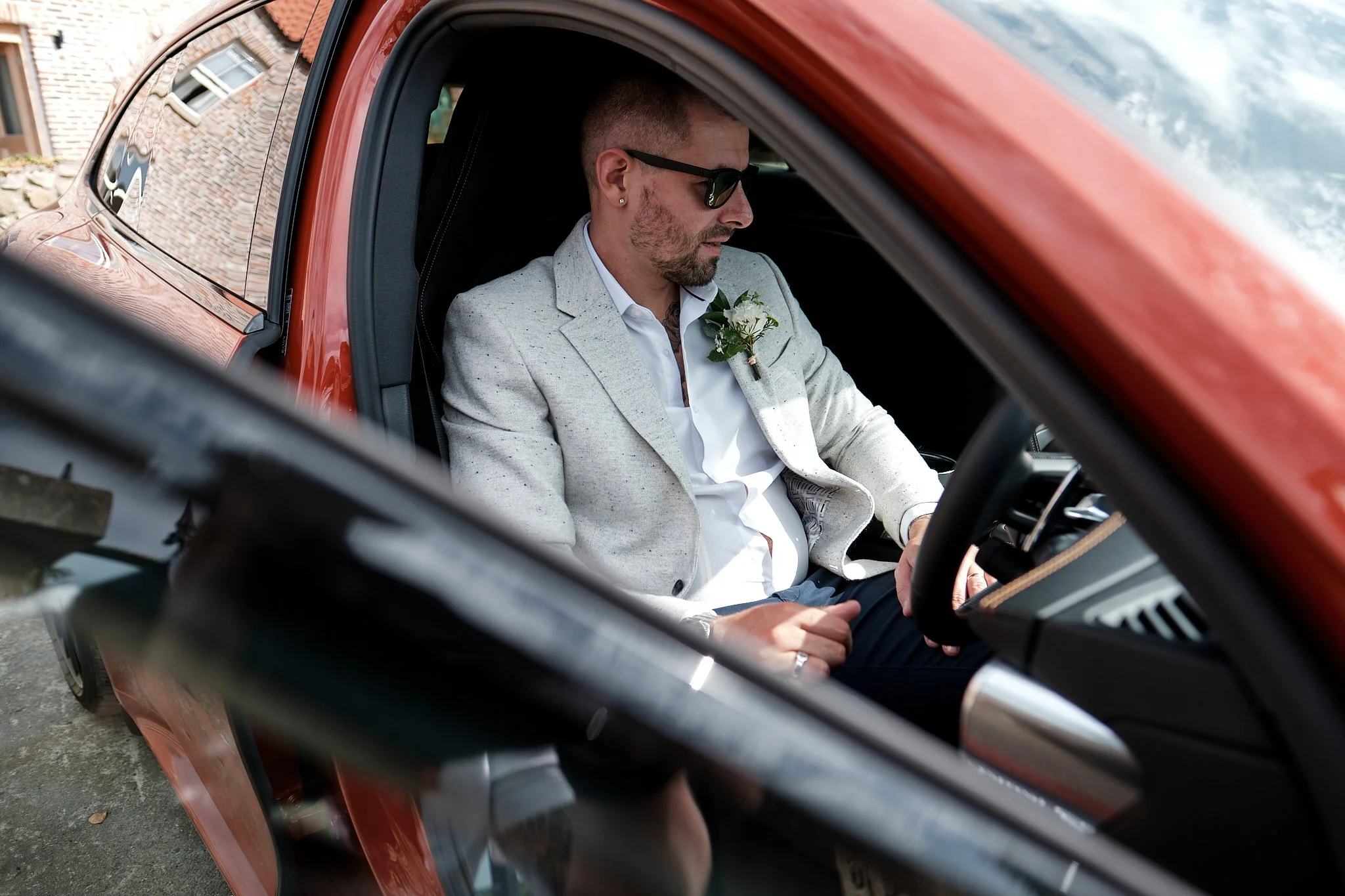 A man with sunglasses and a beard sitting in the driver’s seat of a red vehicle, dressed in a light-colored blazer and white shirt, looking down at something in his lap.