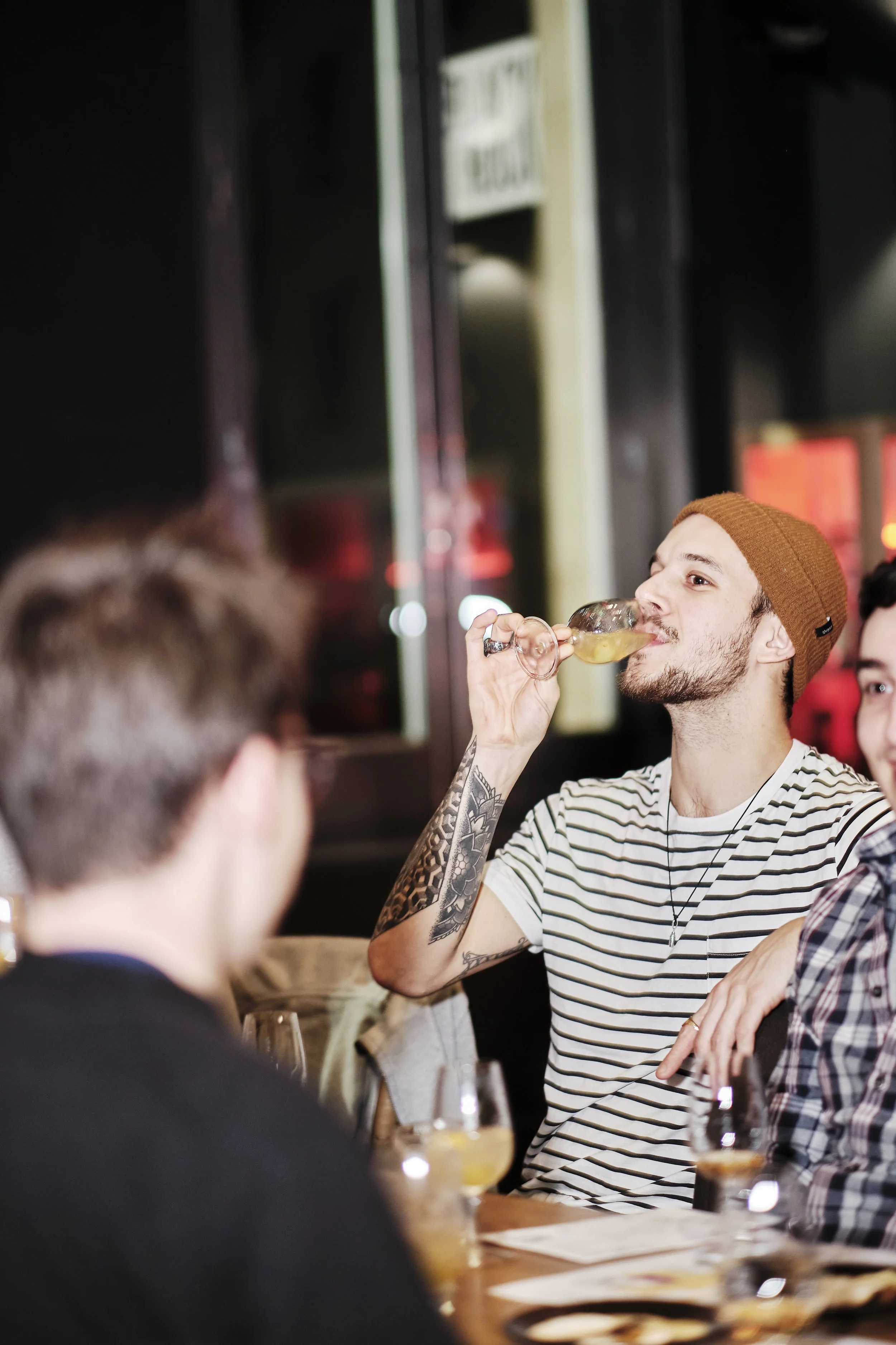 A man wearing a brown beanie and striped shirt is drinking a cocktail at a social gathering, seated at a table with other people.
