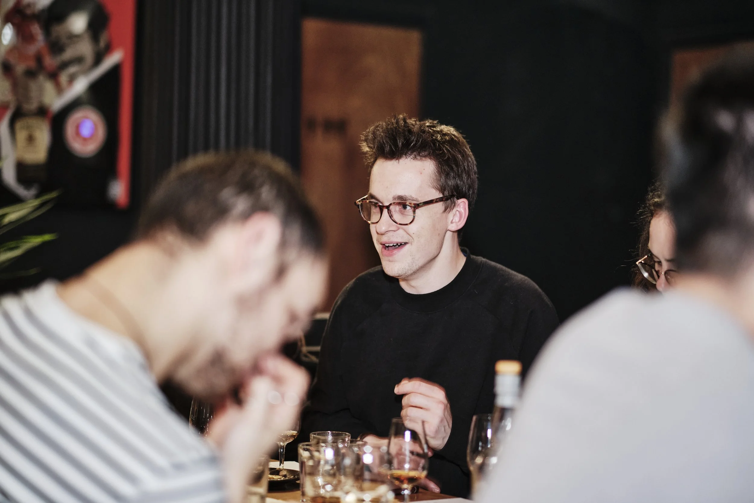 A group of people sitting at a table, with a man in the center wearing glasses and a black sweater, smiling and engaging in conversation, in a dimly lit indoor setting.