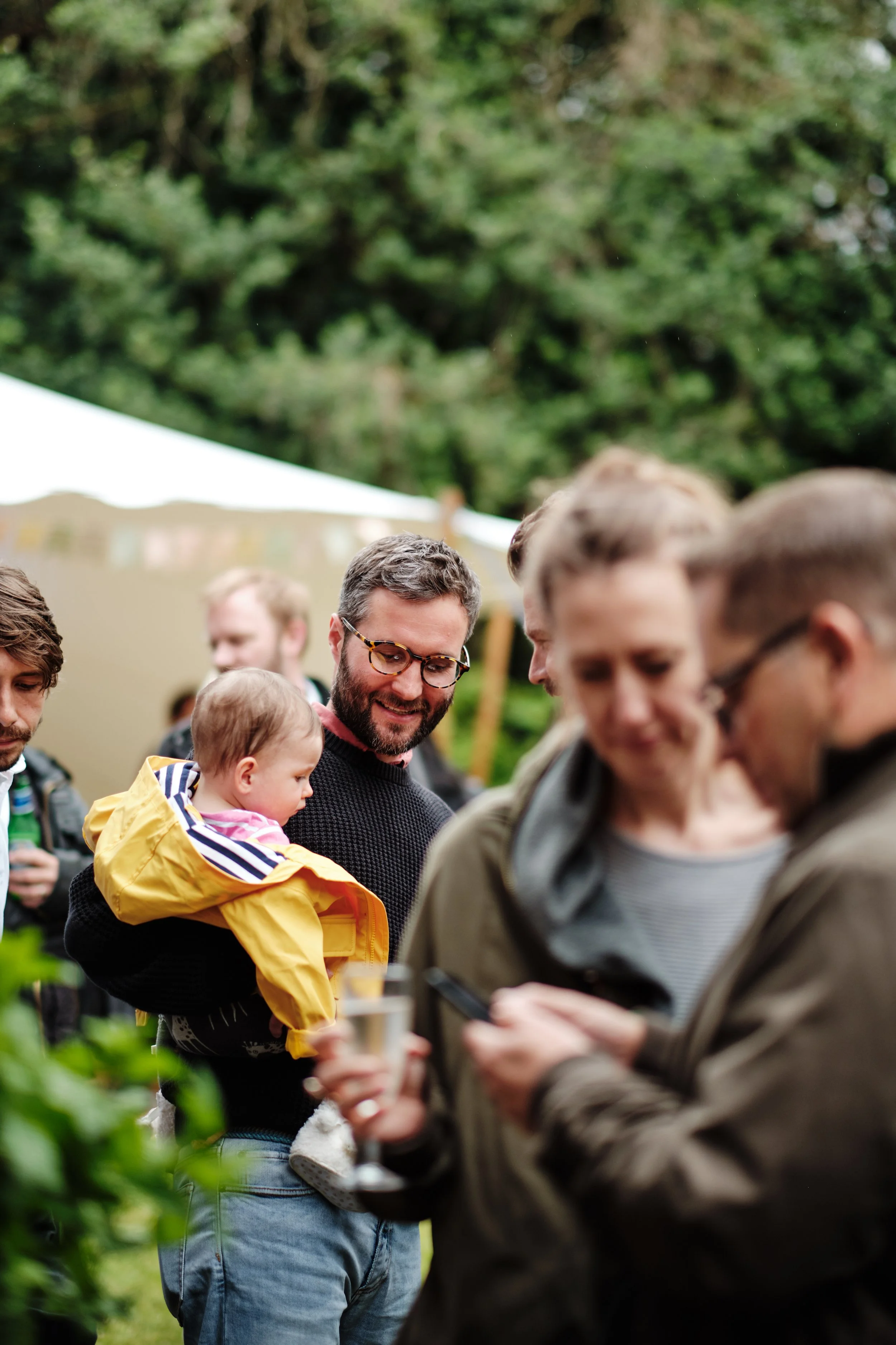 Group of people gathered outdoors at an event, with a man holding a baby in a yellow jacket, smiling, and others looking at their phones.