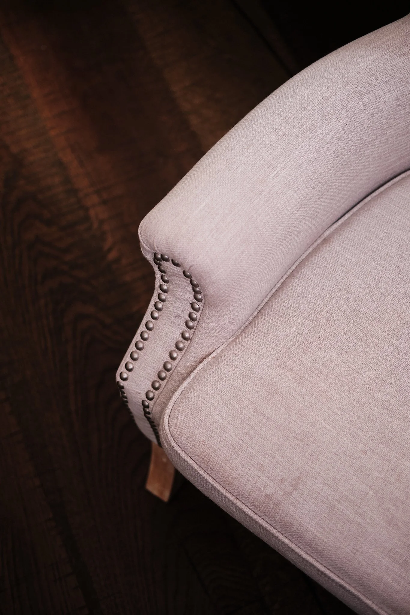 Close-up of a light pink upholstered armchair with nailhead trim on the armrest, situated on a dark wooden floor.