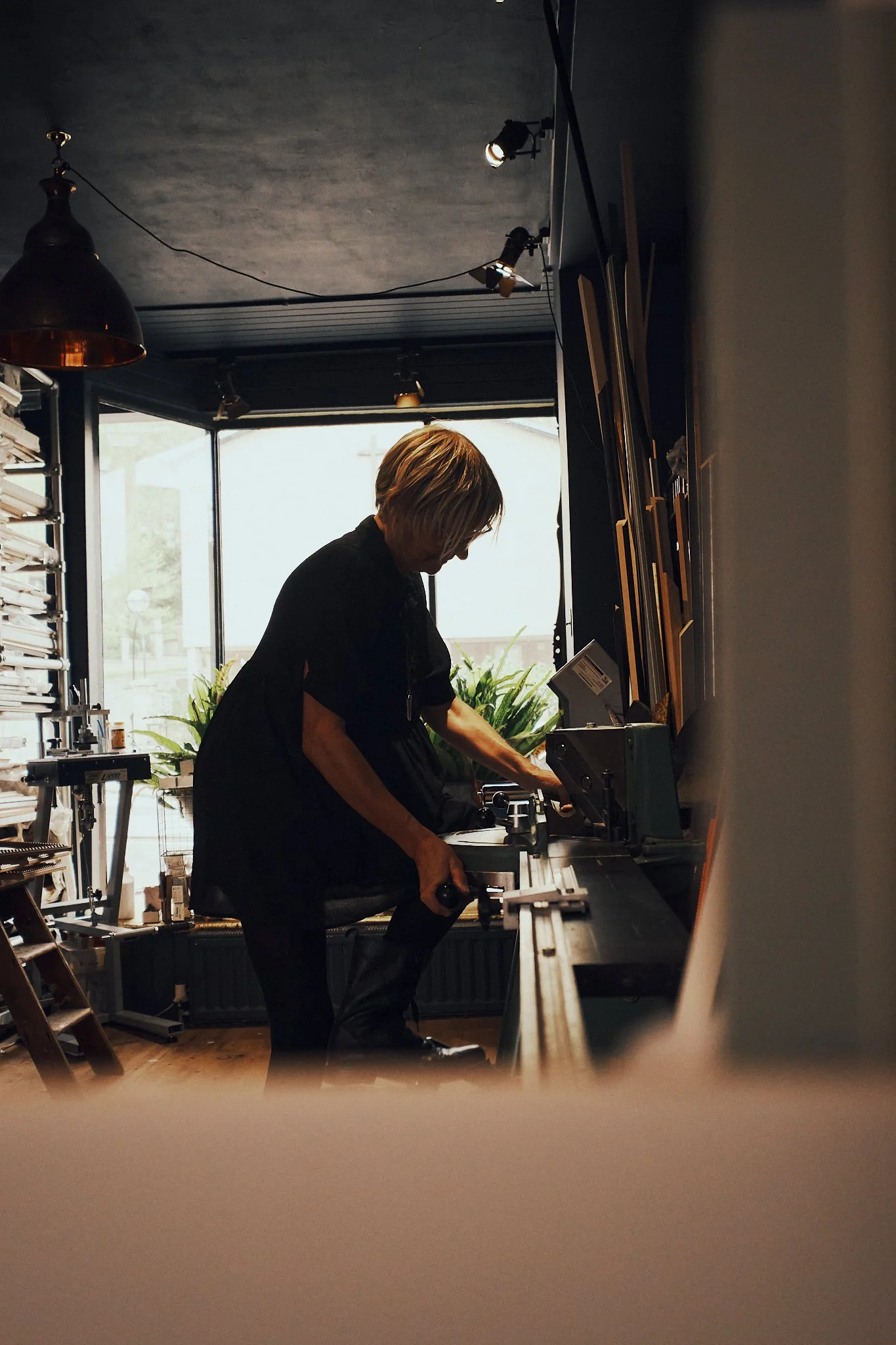 A person with short blond hair working with art supplies in a dimly lit art studio near a large window.