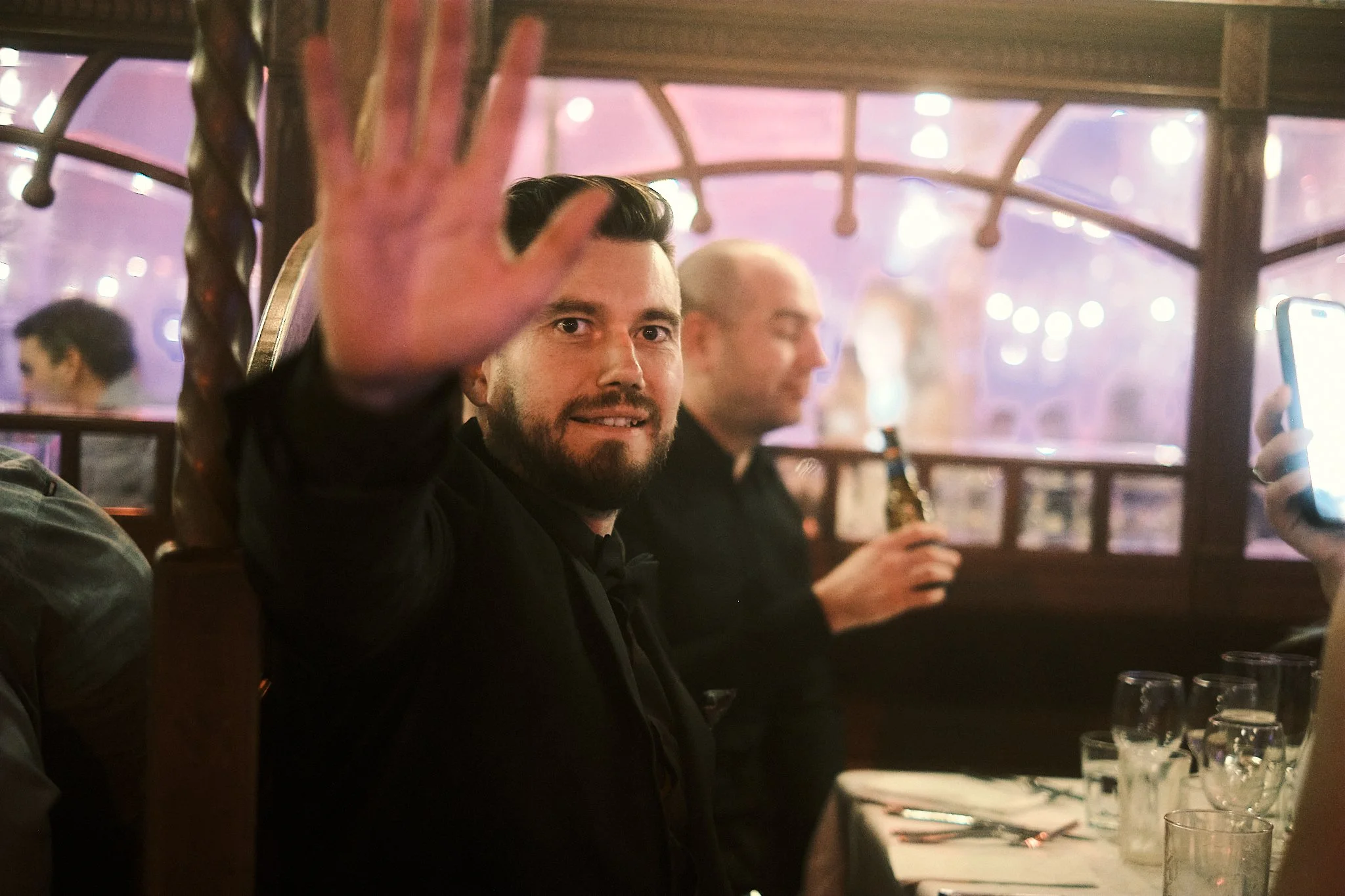 A man with a beard and dark hair, wearing a black jacket, smiling and waving at the camera in a restaurant. Another man in the background is holding a beer bottle, with glasses and tableware visible on the table.