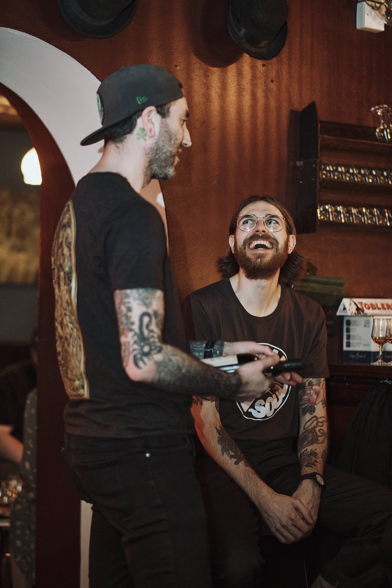 Two men with tattoos and glasses smiling and talking in a bar.