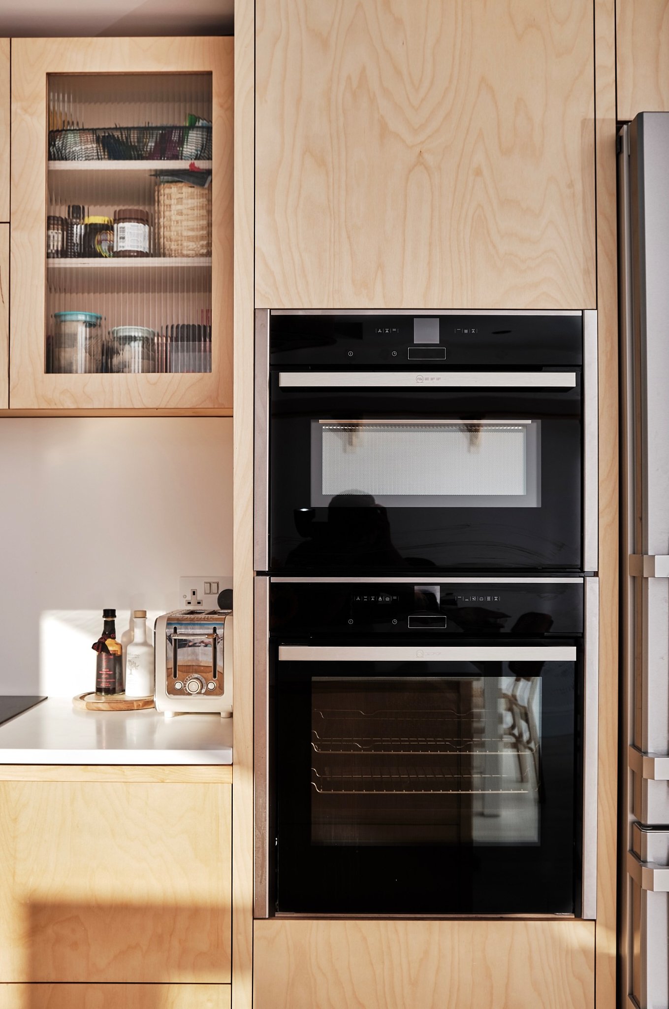 A kitchen with built-in black double oven, a shelf with jars and containers, a toaster on the countertop, and part of a refrigerator on the right.