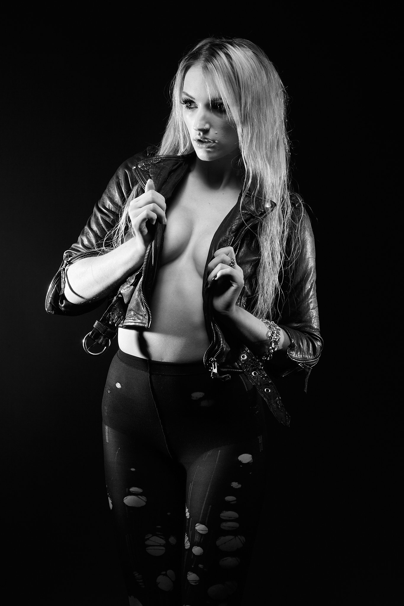 Black and white photo of a young woman with long, wavy hair, wearing a leather jacket partially open, revealing her chest, and ripped leggings. She has a nose piercing and a bracelet on her wrist, standing against a dark background.