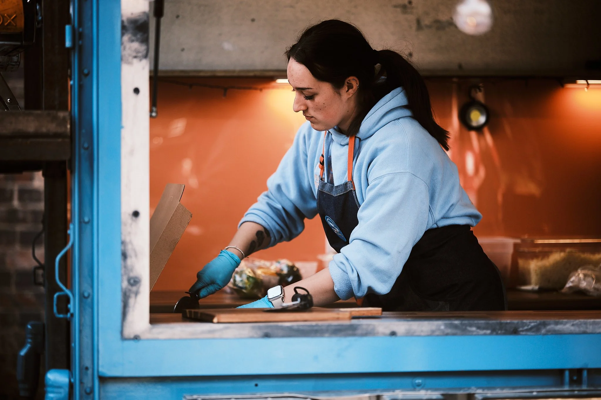 A woman wearing a blue hoodie, black apron, and blue gloves is preparing food in a kitchen or food truck.
