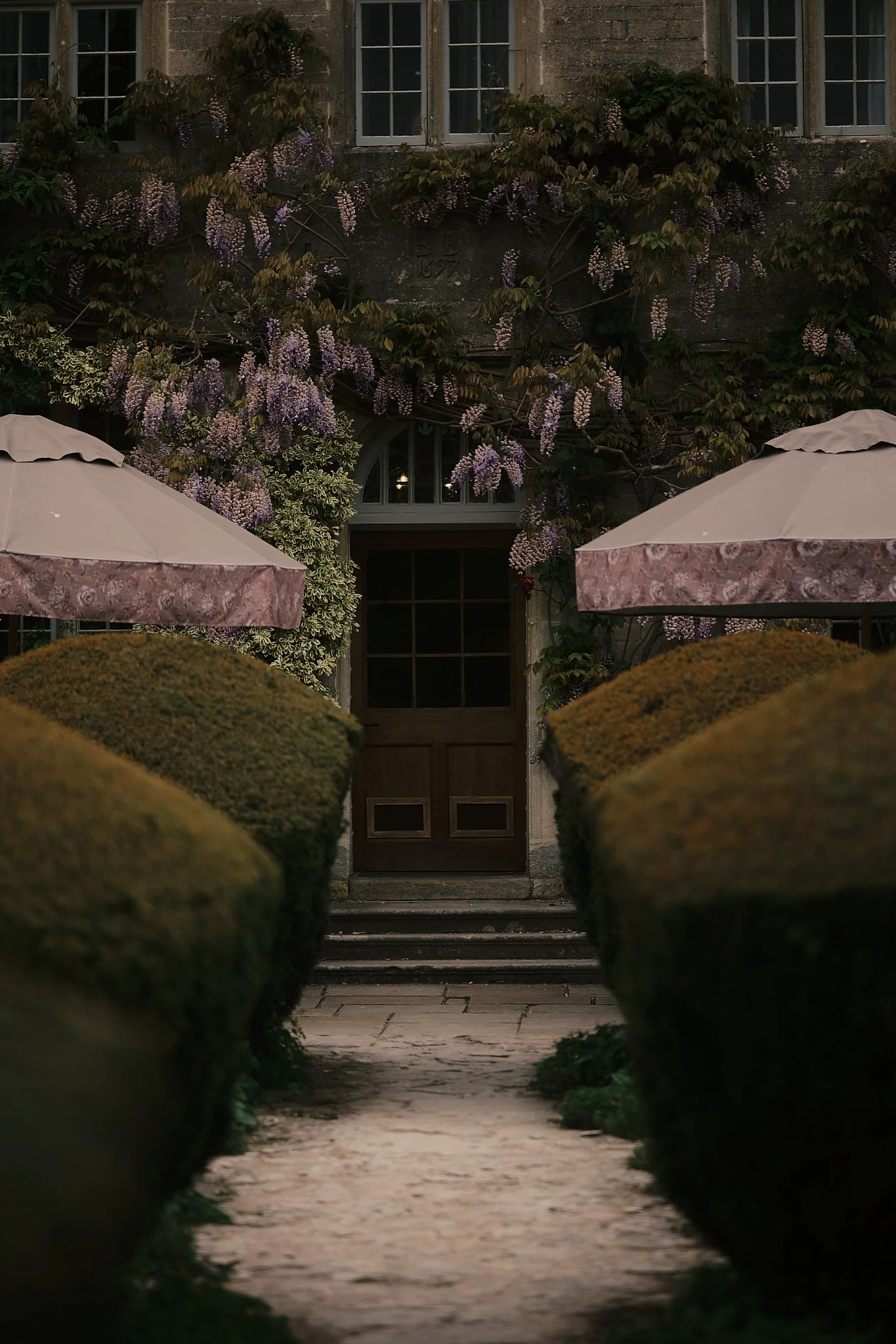 A stone pathway lined with trimmed bushes leading to a wooden door at an old building. The building is covered with purple flowering vines and has two pink patio umbrellas on either side of the pathway. Hotel interior photography at THE PIG-in the Co