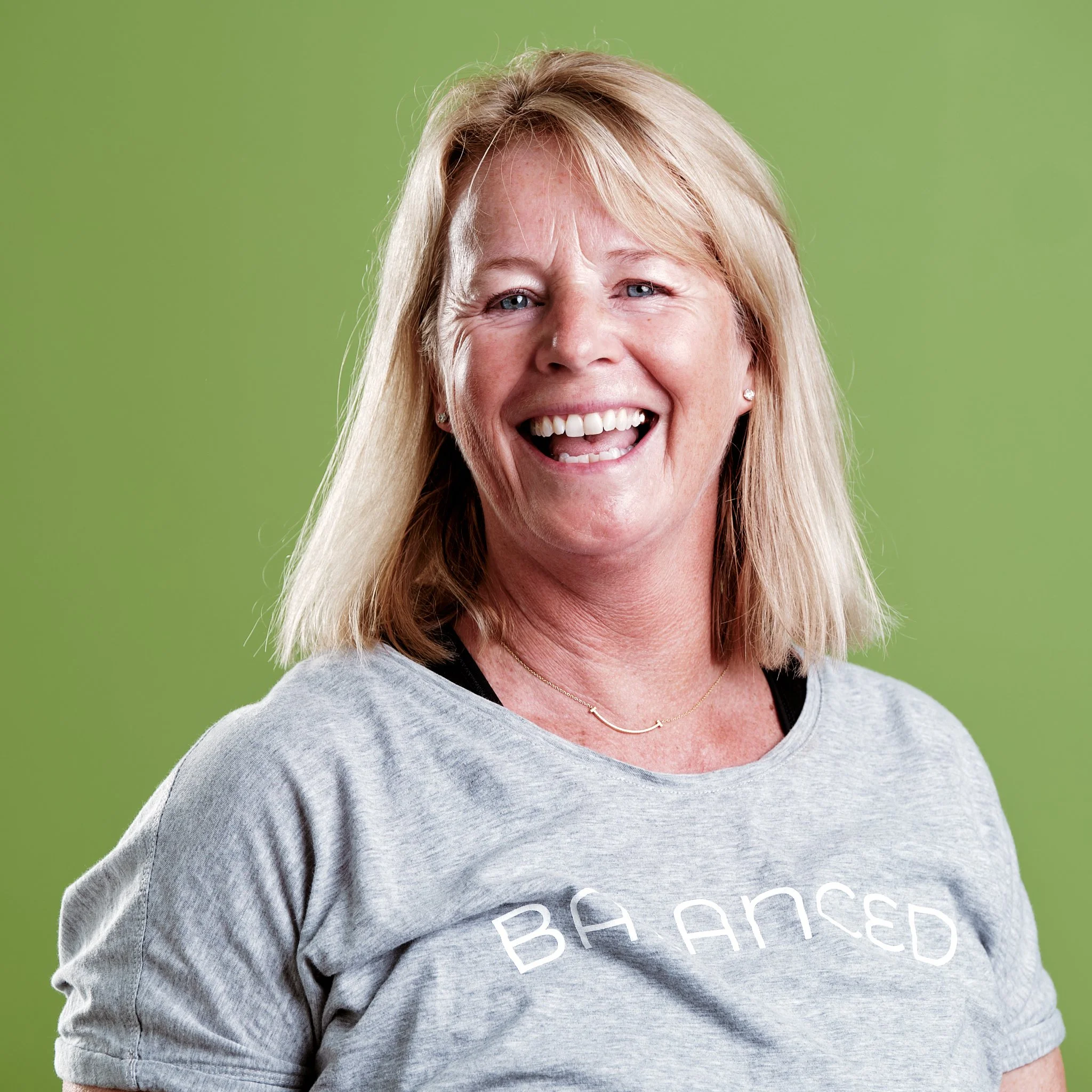 A woman with blonde hair smiling and laughing, wearing a gray T-shirt with partially visible white text, and a black top underneath, against a green background.