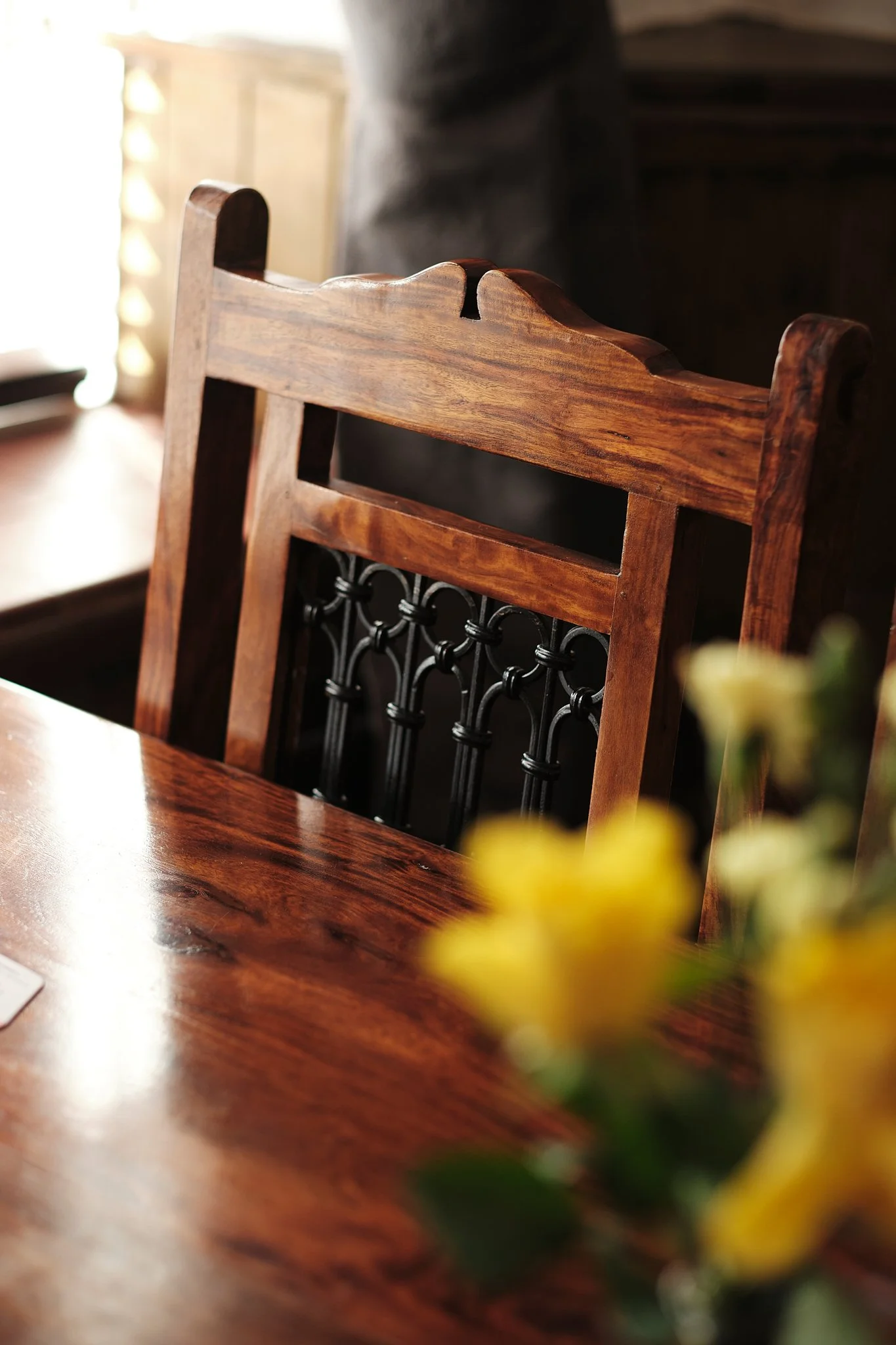 Close-up of a wooden dining chair with black metal decorative back, part of a wooden dining table, with a blurred bouquet of yellow flowers in the foreground.