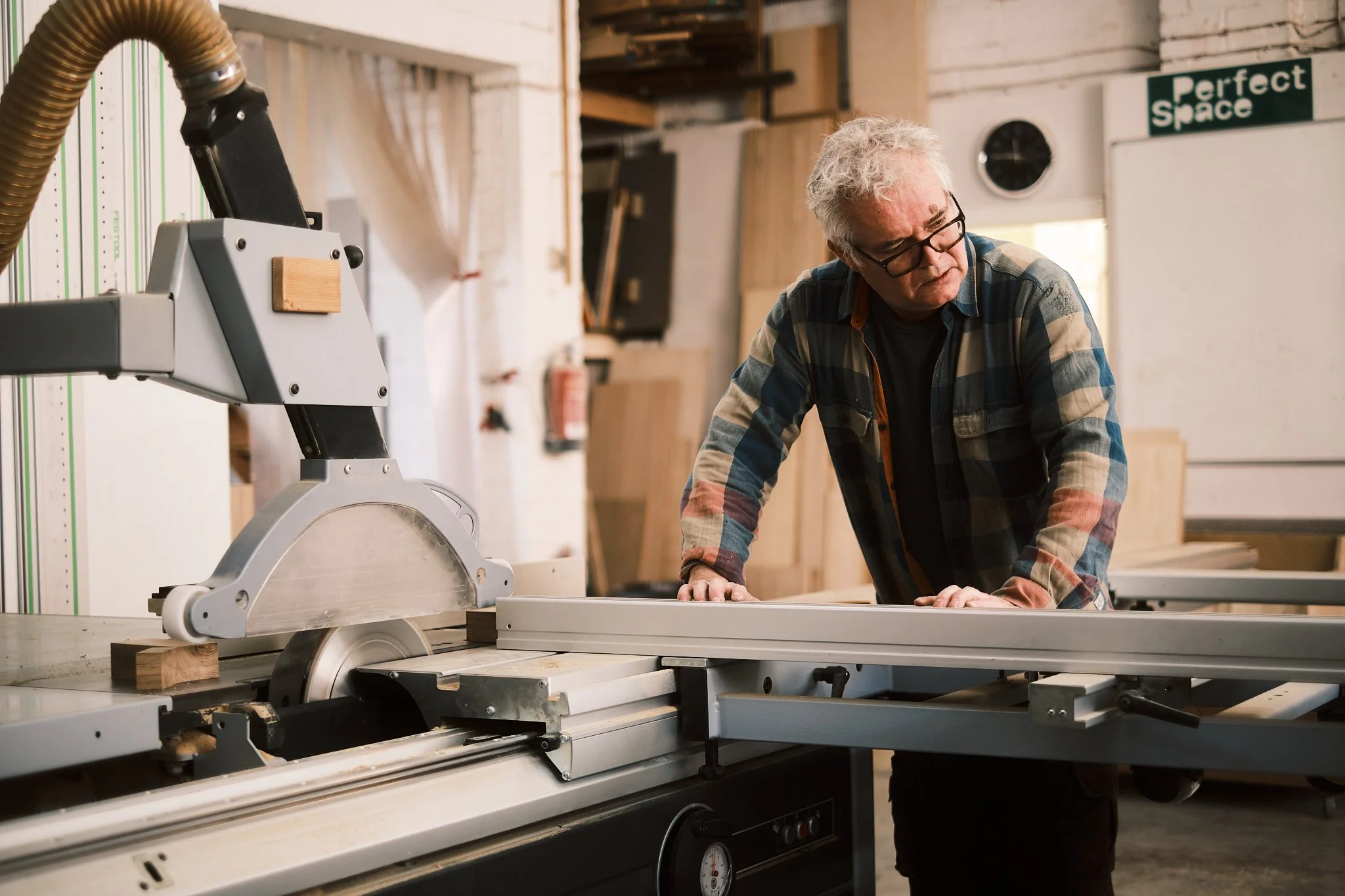 An older man with glasses and a plaid shirt operating a woodworking table saw in a workshop.