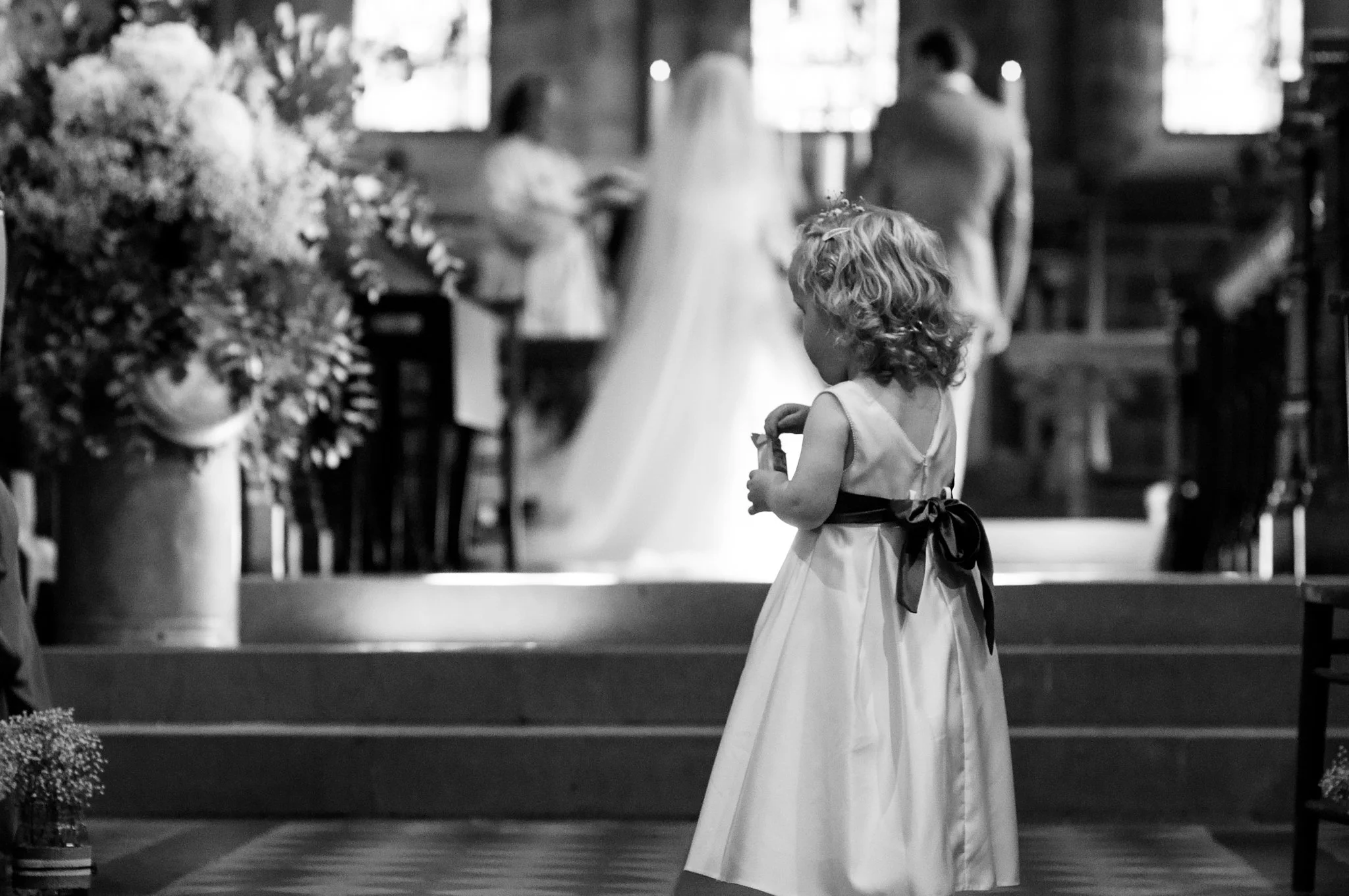A young girl with curly hair wearing a dress with a bow stands indoors, holding a small object, in front of a church altar or similar setting with blurred guests in the background.
