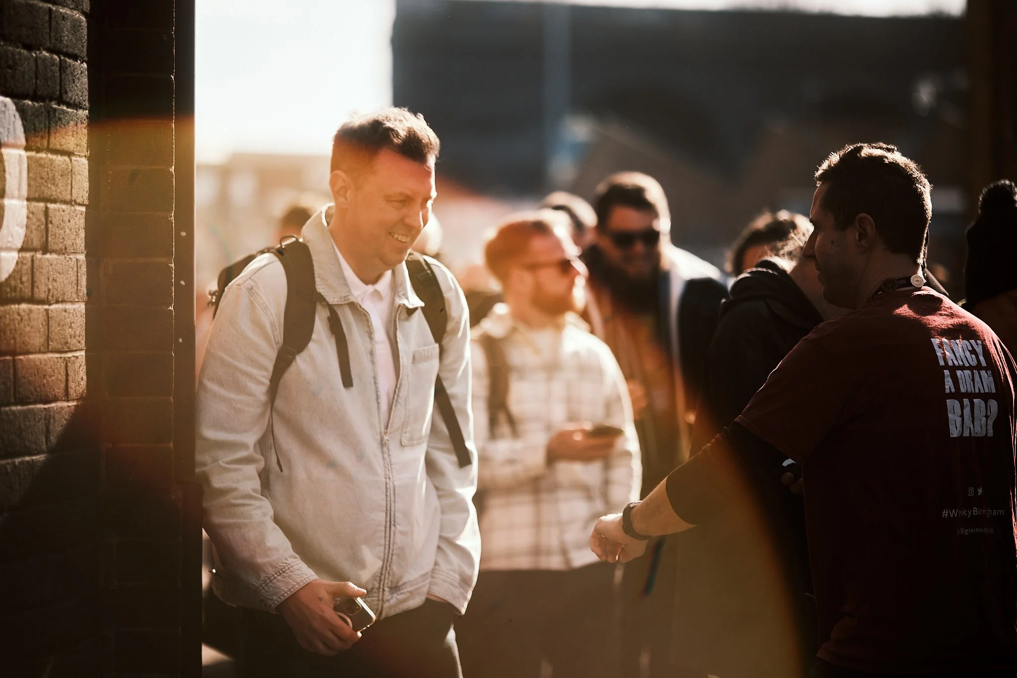 A group of people standing outside in sunny weather, with one person smiling and holding a camera, and others talking and using their phones.