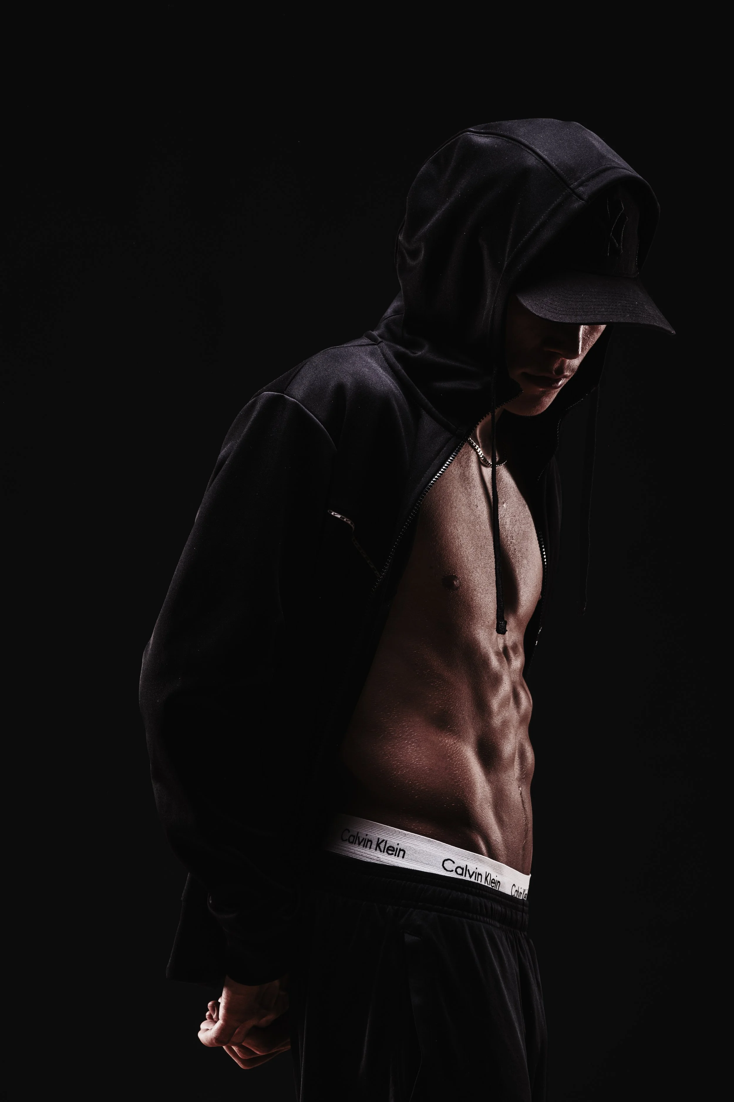A shirtless man wearing a black hoodie and cap, with a Calvin Klein waistband visible, stands against a dark background.