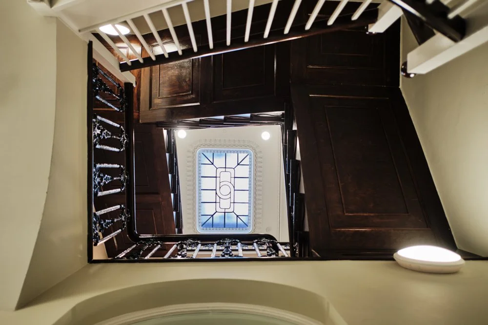 Looking down a staircase with dark wood handrails, balusters, and panels, and a decorative stained glass ceiling window at the top.