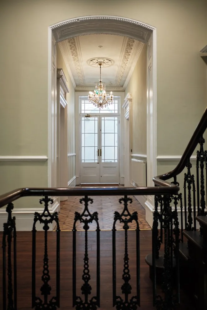 An elegant foyer with a staircase, chandelier, and a front door with glass panes, featuring ornate crown molding and trim.