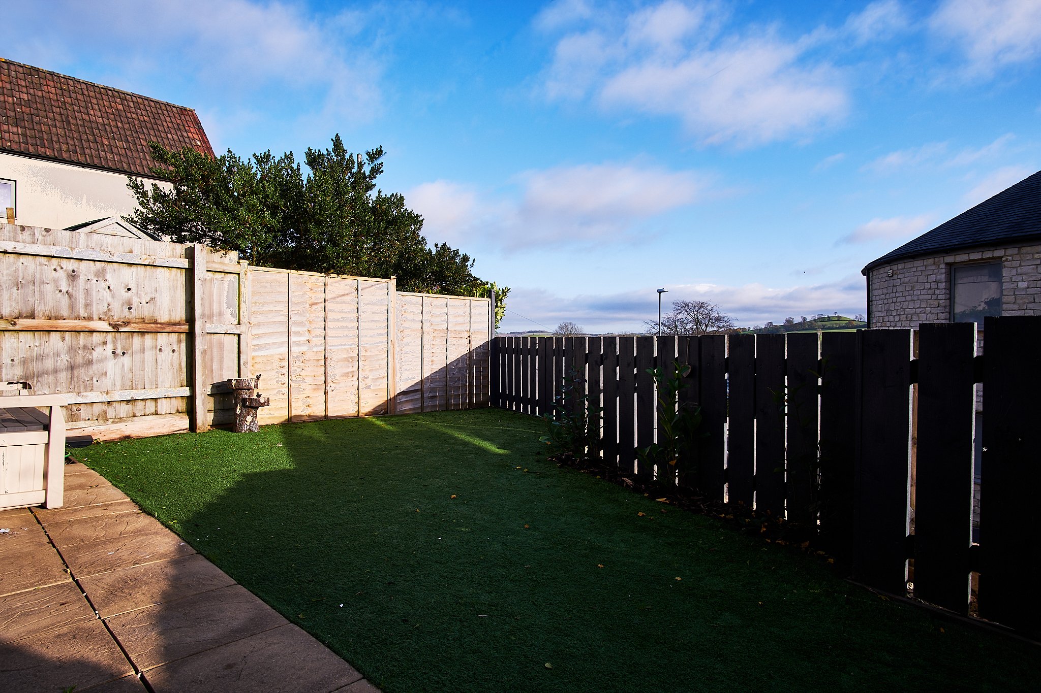 A backyard with a lawn, a wooden fence, and a house with a tiled roof in the background under a partly cloudy sky.