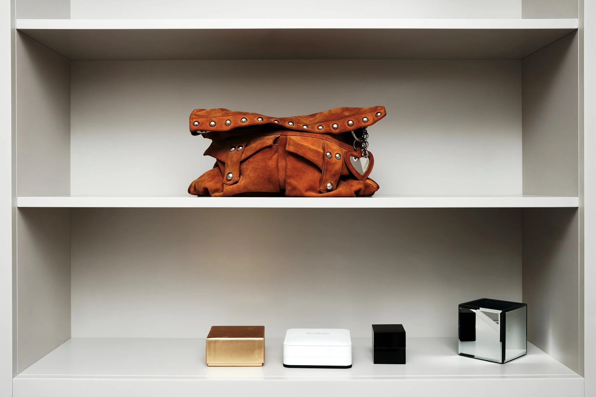 Empty white shelf unit with a brown leather handbag with metal studs on the upper shelf, and four small boxes of different colors and sizes on the lower shelf.