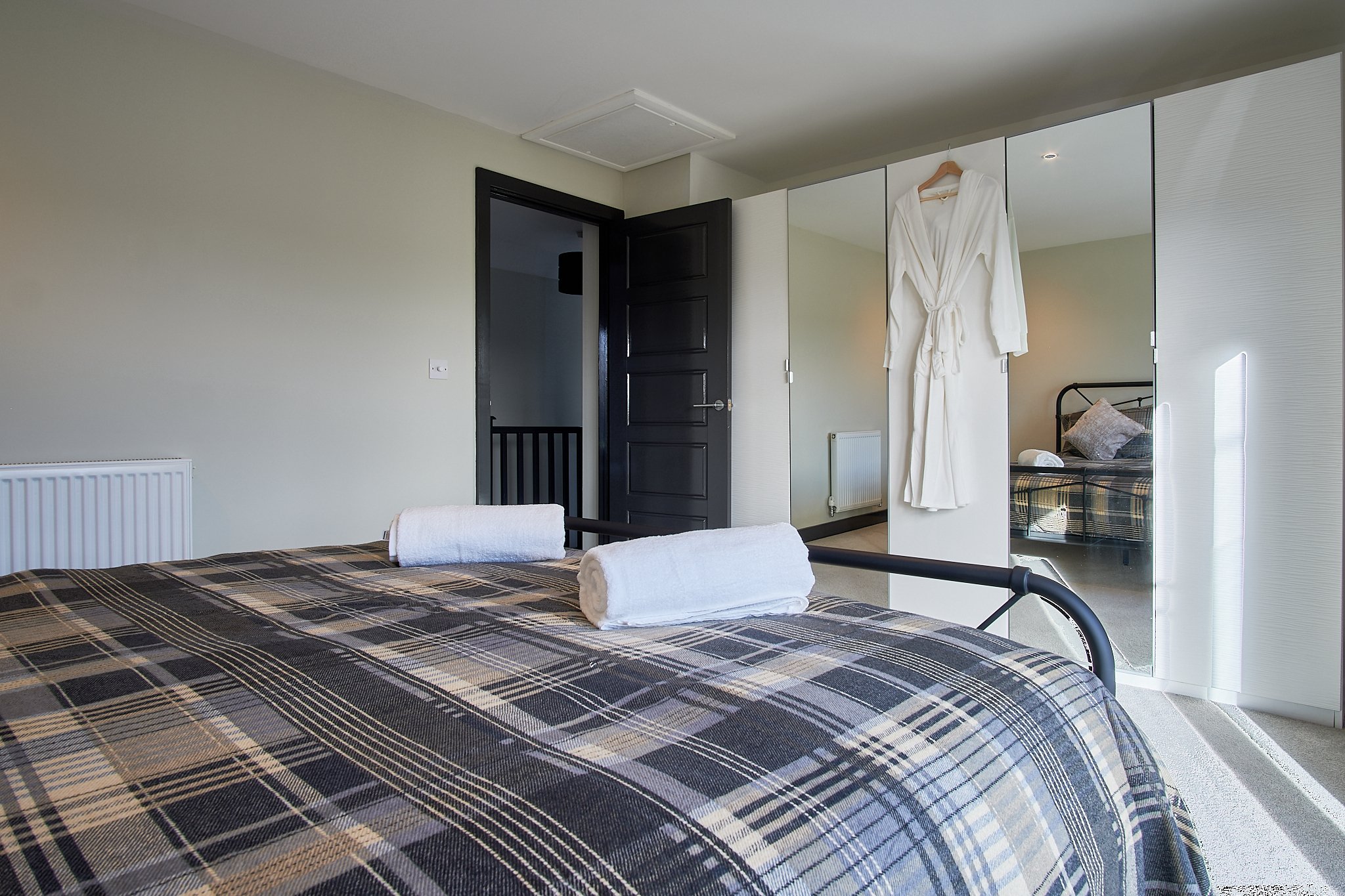 A bedroom with a bed covered in a plaid blanket and two rolled white towels on top. There is a wardrobe with a white robe hanging on it and a mirror. The room has an open door leading to a hallway and a radiator on the wall.