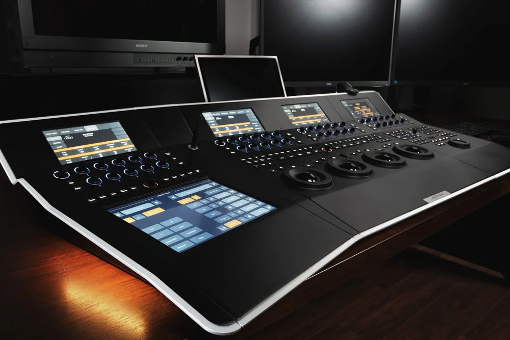 A professional DJ mixing console with multiple screens, knobs, and sliders, placed on a wooden table with computer monitors in the background.