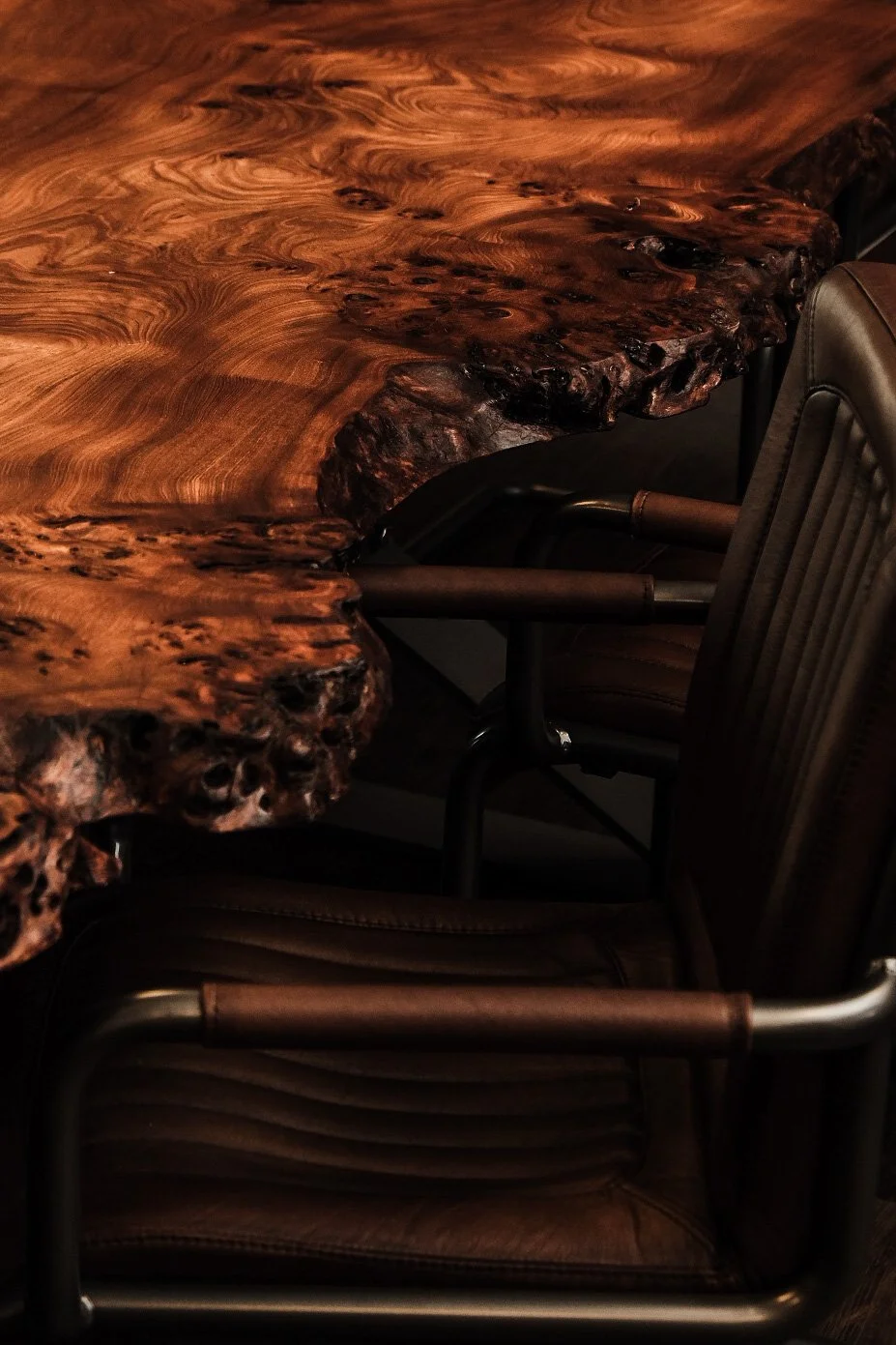 Close-up of a large, polished wooden conference table with a live edge, accompanied by leather office chairs.